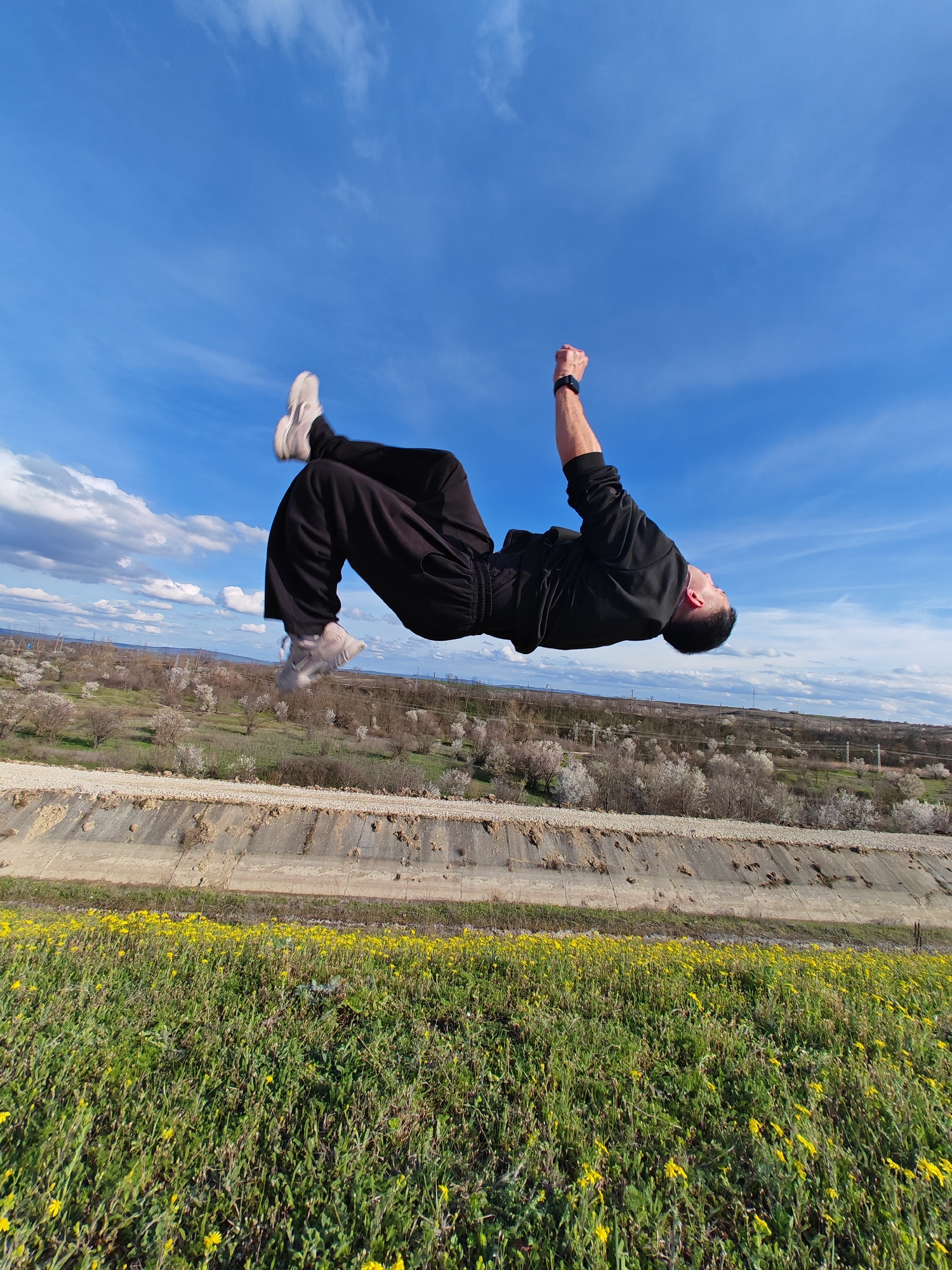 A man in mid-air performs a backflip. He wears black clothing, including a long-sleeved shirt and pants, and light-colored sneakers. His right fist is clenched. The background consists of a clear blue sky with scattered white clouds above a landscape of green fields with yellow flowers and what appears to be a concrete irrigation canal. Distant trees, some in bloom, are also visible. The location is likely near Shumen, Bulgaria, based on the landscape features and the presence of orchards and agricultural lands in that area.