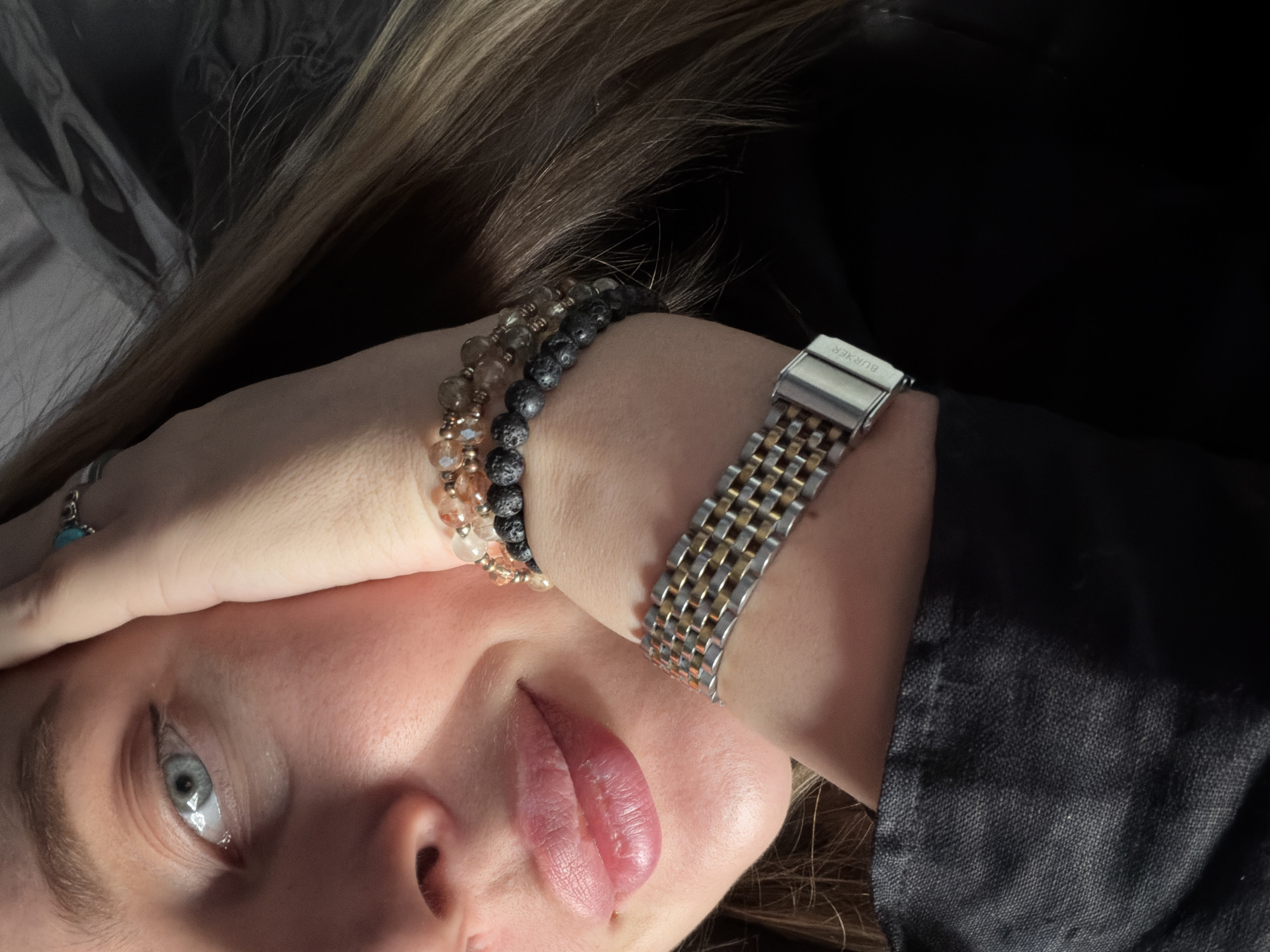 This close-up, high-angle image captures a partial view of a person, likely female, lying down with their head tilted. The lower half of their face is visible, featuring a clear blue-grey eye looking upwards, a nose, and slightly chapped lips. A hand rests across the forehead, partially obscuring the upper face. Long, light brown hair cascades around the head, blending into a dark background.
On the visible wrist, the person wears two beaded bracelets: one with light, translucent beads and another with darker, textured beads, possibly lava stone. A silver-toned ring with a light blue, turquoise-like stone is on the ring finger of the hand covering the forehead. A metal watch with a two-tone (silver and gold) jubilee-style bracelet is worn on the opposite wrist, with the clasp clearly displaying the brand name "BURKER". The person is wearing dark, textured clothing, possibly a black or dark grey shirt or jacket, visible on the right side of the frame. The background is indistinct and dark, suggesting an indoor or dimly lit setting. The overall mood is contemplative or relaxed, with the direct gaze of the eye being a notable detail. The location context provided for this media file is Moscow, Russia.