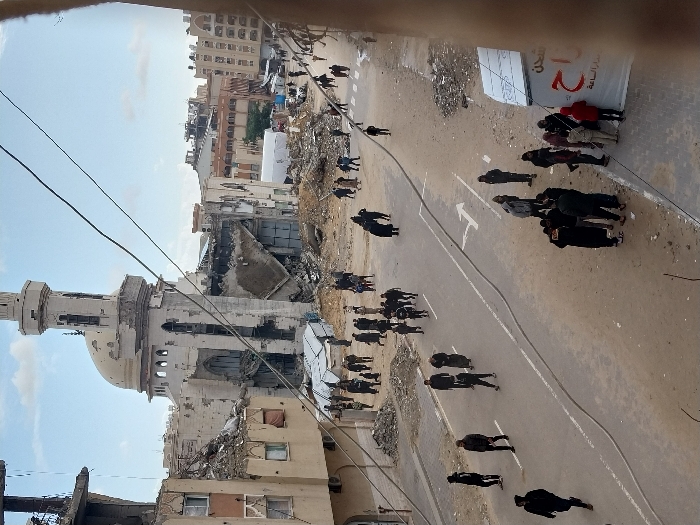 This overhead image depicts a scene of extensive urban destruction in Khan Yunis, Palestinian Territory. The central feature is a severely damaged mosque, identifiable by its minaret and dome, both exhibiting visible structural compromise, including large holes in the dome and significant facade damage. The minaret's upper section shows exposed internal structure.
Surrounding the mosque, multi-story buildings are in various states of collapse and disrepair, with walls missing, facades blown out, and interiors exposed. Piles of rubble, concrete, and twisted rebar are scattered across the ground, particularly around the damaged structures.
A paved street, marked with a white centerline and a "turn right" arrow, runs through the foreground and midground. It is partially covered in dirt and debris. Numerous individuals, mostly adults, are present on the street and in the surrounding areas. They are dressed in civilian clothing and are seen walking individually or in small groups. Some are congregated on the sidewalk near a white tent-like structure with red Arabic text on a banner.
The overall environment conveys a sense of post-destruction activity, with people moving through a heavily damaged urban landscape under an overcast sky. Overhead utility lines are visible crisscrossing the upper portion of the image.