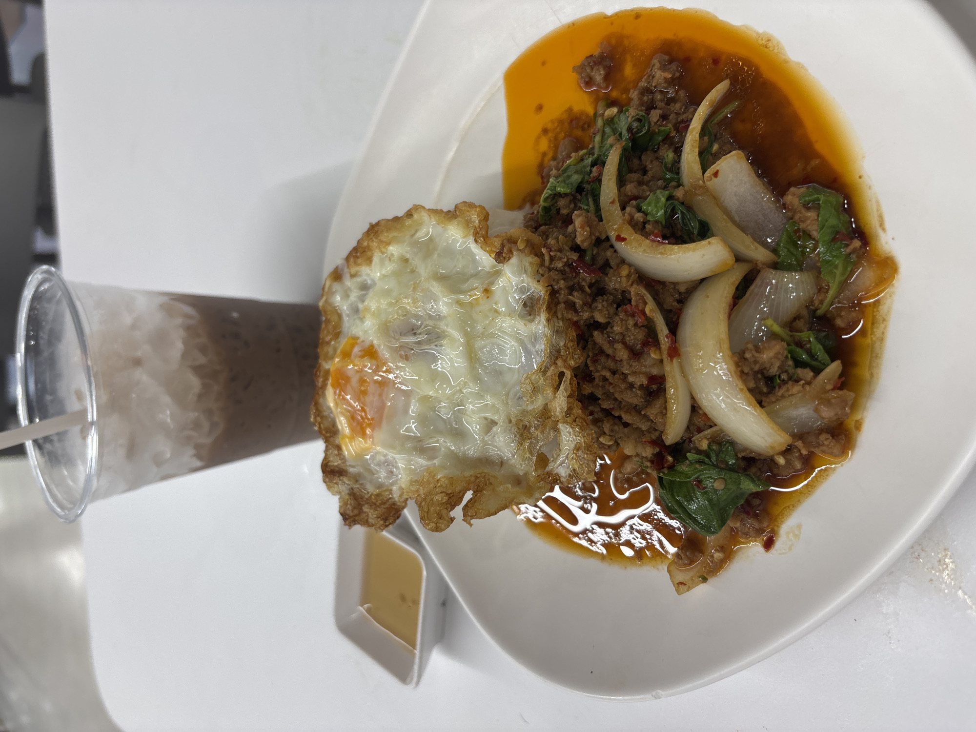 A plate of Pad Kra Pao Moo is presented on a white plate, featuring stir-fried minced pork with Thai basil and onions, served with a fried egg. A small container of sauce rests near the egg. Beside the plate is a plastic cup containing a beverage, possibly chocolate milk, with a straw inside. The setting is a white table in บ้านบางรักษ์, Thailand.