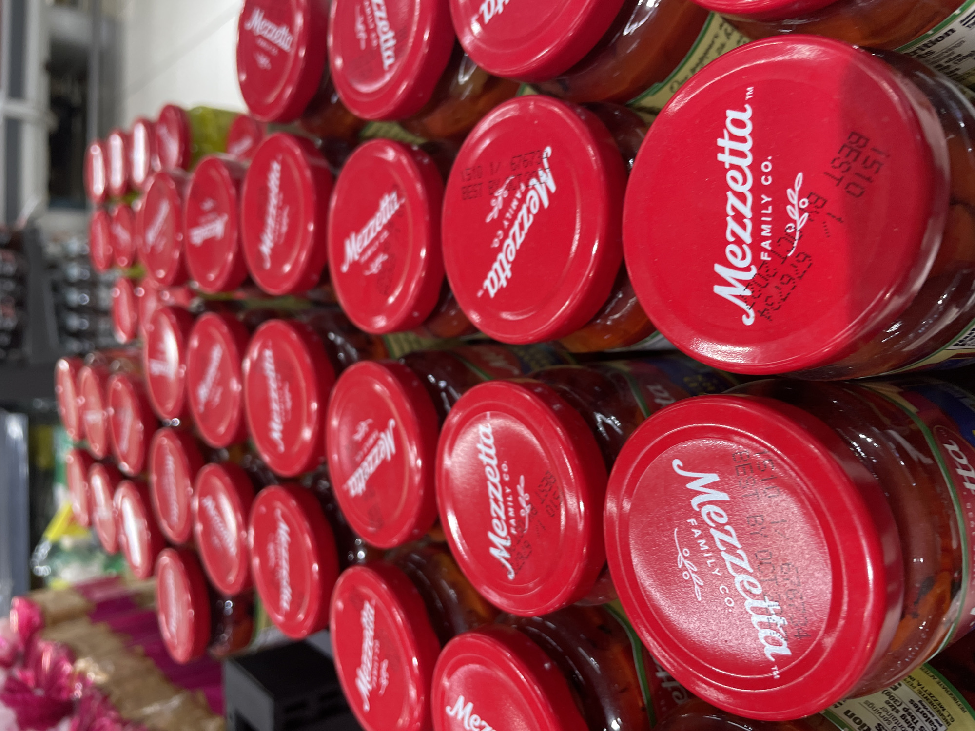 The image shows a close-up, eye-level view of multiple jars of Mezzetta brand food products on display, likely in a grocery store. The jars have bright red lids with the "Mezzetta FAMILY CO." logo prominently displayed in white text. Some lids also show stamped best-by dates and product codes. The products inside the jars are visible, appearing to be various types of preserved peppers or similar items in liquid. The background is blurred, suggesting a retail environment with other products and shelving. The lighting is bright and appears to be artificial, consistent with indoor retail lighting. The perspective is focused on the arrangement of the jars, with several rows visible, extending into the background. The scene is static and does not depict any action or people. The specific location in Davao City, Philippines, cannot be determined from the visual information provided in the image itself.