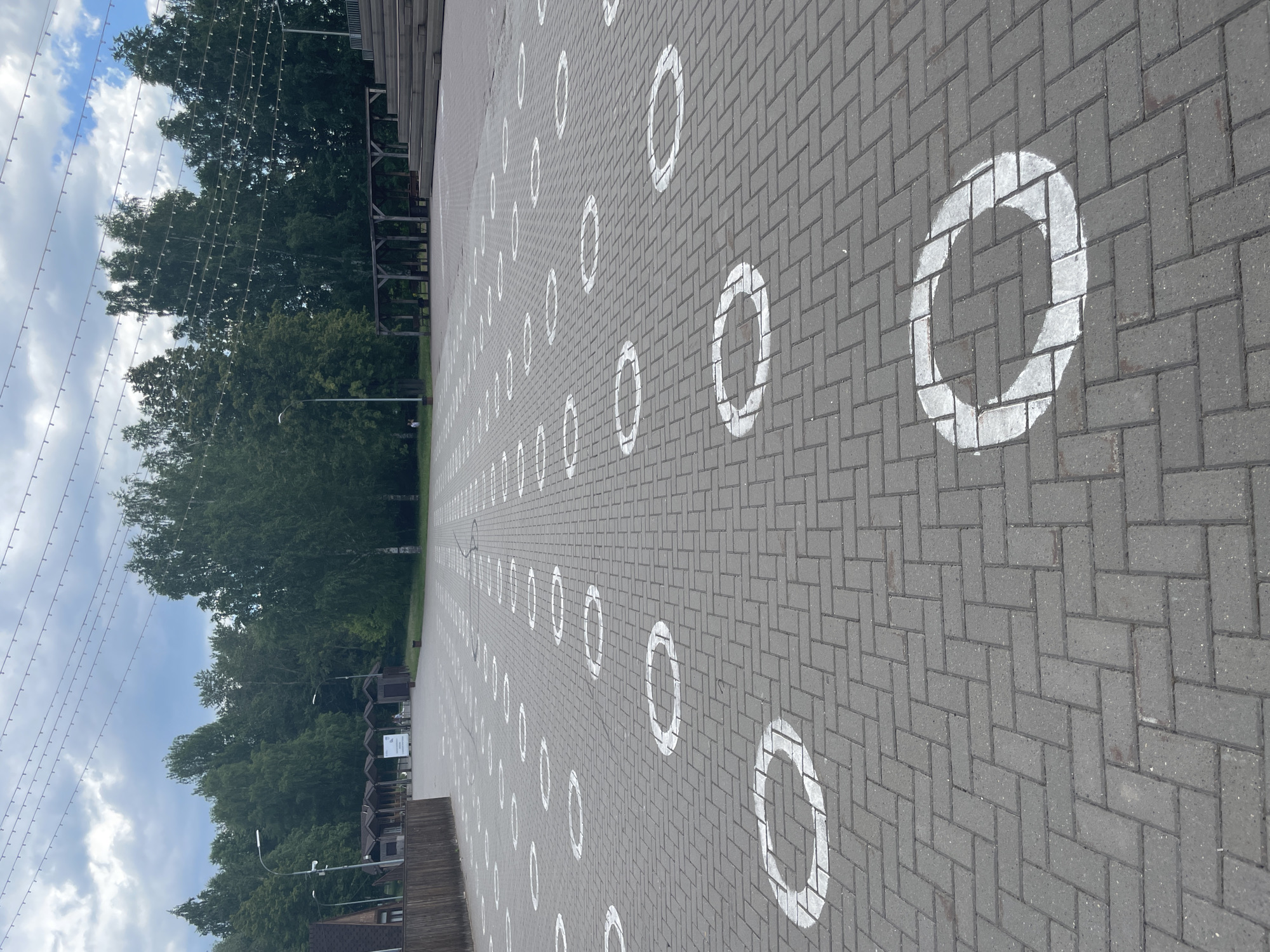 The image shows an outdoor paved area with a herringbone pattern of gray brick pavers. White circular markings, approximately one meter in diameter, are painted on the pavers, spaced out in a grid-like pattern. These markings appear to be social distancing circles. In the background, there are trees, some covered walkways or gazebos, and a tiered seating area that resembles an outdoor amphitheater. Overhead, strings of small lights are strung across the scene, suggesting a public gathering space. The sky is partly cloudy. The location is identified as the Central Park of Culture and Recreation in Izhevsk, Russia. No people are clearly visible in the image, though faint figures may be present in the distance.