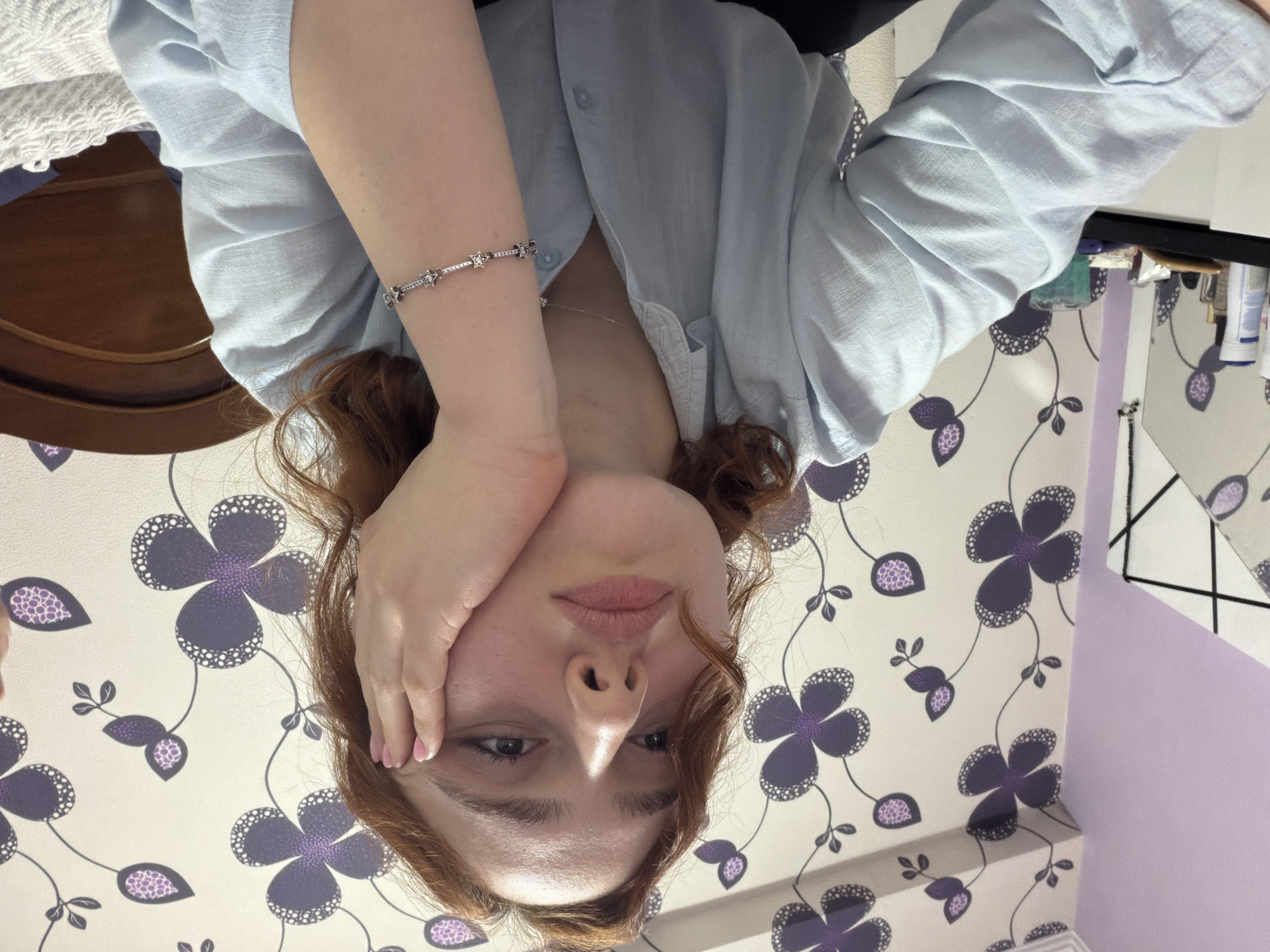 A woman with red hair and pale skin is pictured, facing the camera. She is wearing a light blue button-down shirt and a silver bracelet. Her left hand is touching her face near her cheek. The background is a patterned wallpaper with purple and white flowers. A shelf with toiletries is visible in the upper right corner. The image is taken at an angle, suggesting she might be lying down. There is no indication of a specific location in Tbilisi, Georgia.