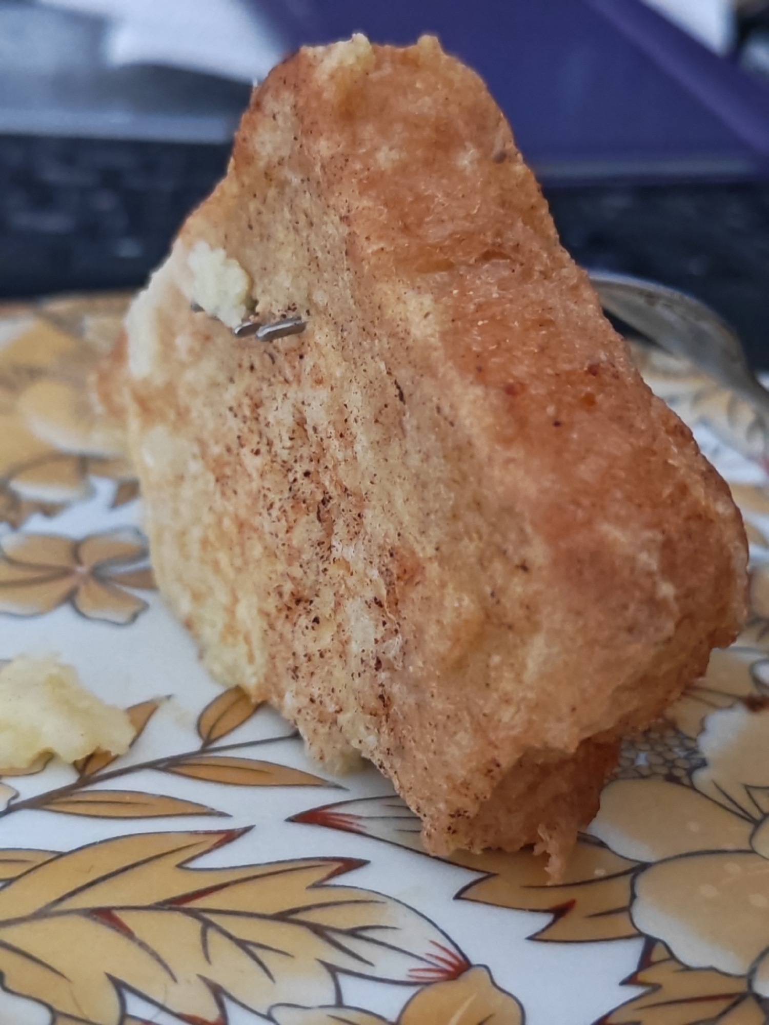 The image shows a close-up of a piece of food, possibly cake or bread, with a few tiny metal fragments embedded in it. The food item is golden brown and appears to be somewhat crumbly. It sits on a plate with a decorative pattern of yellow and brown floral motifs. In the background, out of focus, there is a dark blue object and a blurred surface, likely a table. The setting does not provide any specific indicators of its location within the city of Vanadzor, Armenia, or any other city. There are no people, actions, or interactions depicted.