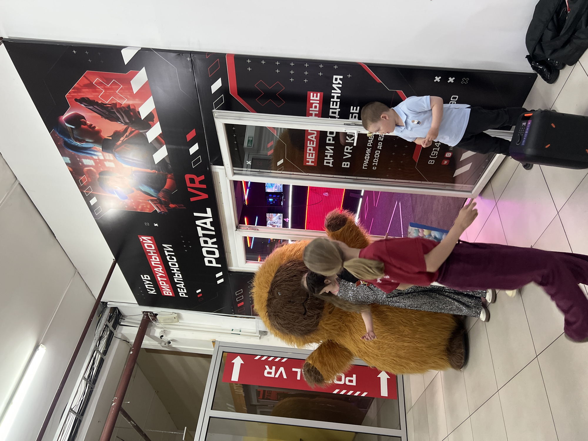 An indoor scene depicts the entrance of a Virtual Reality (VR) club named "Portal VR." The club's facade is decorated with a black and red graphic featuring a person in a VR headset holding a weapon, geometric patterns, and text.
In the foreground, a large, furry, brown mascot, possibly a lion or dog character, stands near the entrance. Two children are interacting with the mascot: a young girl in a sparkly dress is close to its head, and another child, mostly obscured, is also nearby. Standing next to the mascot is a woman with blonde hair, wearing a maroon long-sleeved shirt and matching trousers, looking towards the children. To the right, a young boy in a light blue polo shirt and dark pants stands independently, looking down. A black portable speaker with colorful lights is positioned next to him.
Above the entrance, exposed wiring and security cameras are visible. The floor is tiled.
Visible text on the club's signage includes "КЛУБ ВИРТУАЛЬНОЙ РЕАЛЬНОСТИ" (Club of Virtual Reality) and "PORTAL VR." Promotional text advertises "НЕРЕАЛЬНЫЕ ДНИ РОЖДЕНИЯ В VR КЛУБЕ" (Unreal Birthdays in VR Club). Operating hours are listed as "ГРАФИК РАБОТЫ с 10:00 до 22:00" (Operating hours from 10:00 to 22:00), along with a partially visible phone number "8 (914) 90..." and icons for social media platforms like VK and YouTube. The glass door reflects upward-pointing arrows and "PORTAL VR" in reverse. The overall atmosphere suggests an entertainment venue for children and families.