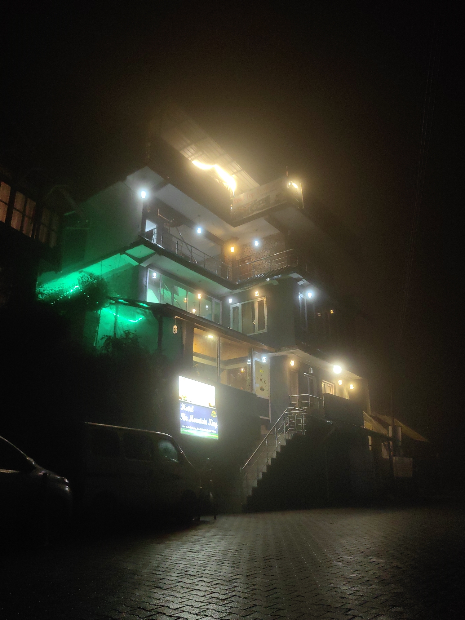 The image captures a nighttime view of the Hotel The Mountain King in Vashisht, India. The hotel is a multi-story building illuminated by various lights, including green lights accentuating the lower facade and warm yellow lights from windows and external fixtures. The building's architecture appears modern with balconies and large windows. In front of the hotel, a paved area, likely a parking lot, is visible, with a portion of a dark-colored van and another vehicle on the left side of the frame. A lit sign for "Hotel The Mountain King" is prominently displayed near the entrance. The scene is characterized by fog or mist, creating a hazy atmosphere and softening the details of the surroundings, contributing to a sense of depth and obscurity. The overall impression is one of a quiet, possibly damp, evening at a hotel situated in a mountainous or hilly region, given the name "The Mountain King".