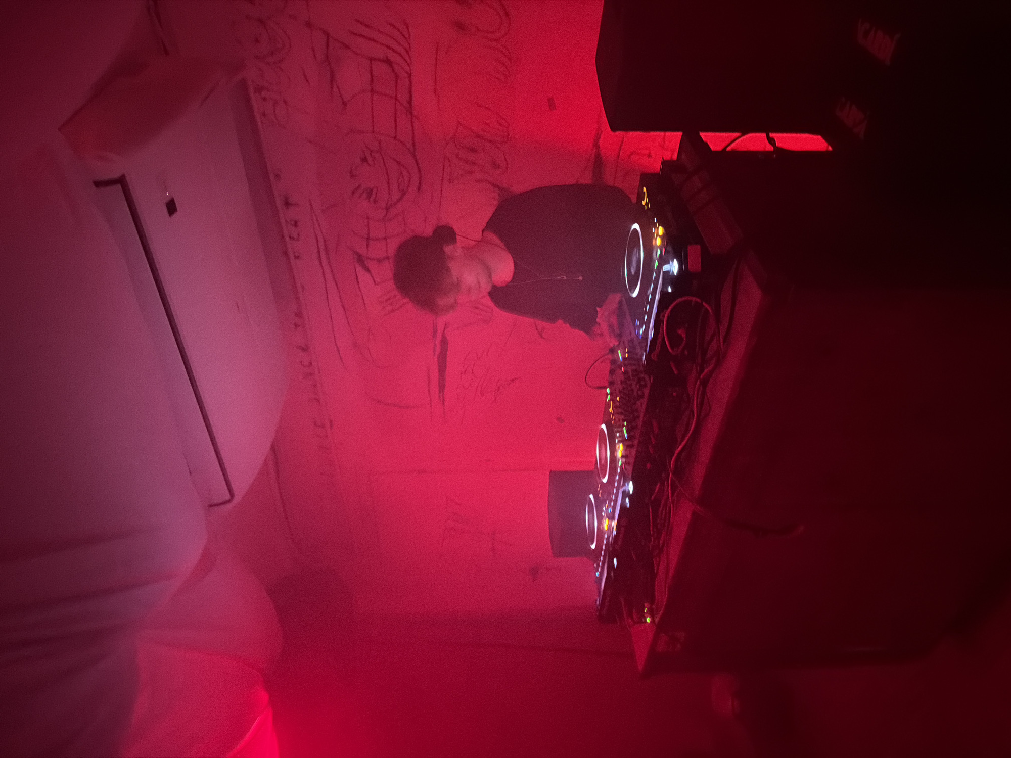 A DJ in a darkened, red-lit room is operating a DJ mixer. The wall behind them is covered in graffiti. An air conditioning unit is mounted on the left side of the frame. Loud speakers are visible on the top right. The city and location are unknown.