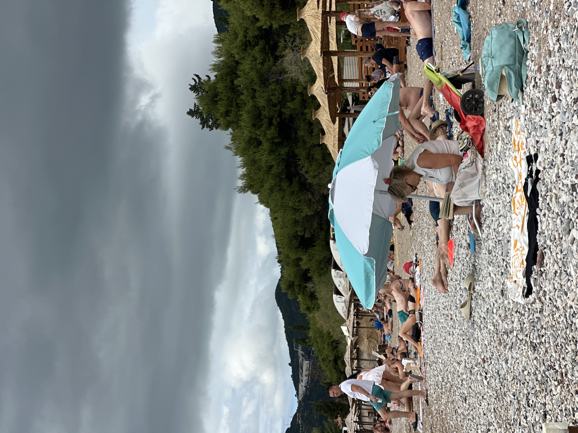 The image depicts a crowded pebble beach under a heavily overcast sky, oriented horizontally despite the vertical framing. In the foreground, a prominent blue and white beach umbrella is open, surrounded by beachgoers. A woman in a white t-shirt bends near a red and green beach trolley and a light blue backpack. Various towels, including a patterned orange and white one and a black one, are spread on the light grey pebbles.
Numerous other people are scattered across the beach, relaxing on towels or loungers, or standing, dressed in swimwear and casual attire. Extending along the midground are several rustic thatch-roofed structures, possibly cabanas or beach bars. Behind these structures, a dense, dark green forest covers a steep mountain that rises dramatically, forming a lush backdrop to the scene.
The sky is dominated by dark, ominous grey clouds, particularly dense on the left side, indicating potentially stormy weather or recent rain. The light is diffused, consistent with a very cloudy day. The overall scene suggests a busy vacation spot, characteristic of a coastal resort. The word "BUDVA" is partially visible on a white bag or t-shirt held by a person in the background, near the thatched structures.