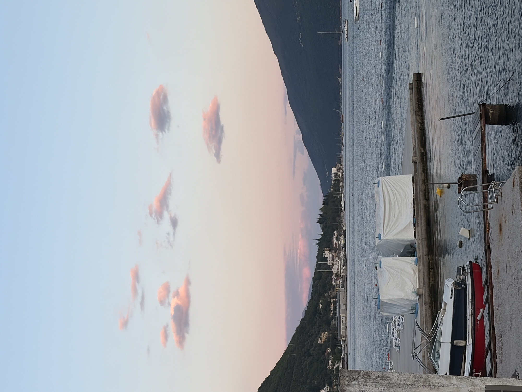 This serene landscape captures a coastal scene at either sunrise or sunset, indicated by the soft, warm light. The sky above is a gradient of light blue, transitioning to pale pink and subtle orange hues near the horizon. Scattered, wispy clouds, reflecting these warm tones, are distributed across the upper part of the sky.
In the mid-ground, a dark, densely forested mountain range rises, its silhouette stretching across the frame. Along the base of these mountains, a town or settlement is visible, characterized by numerous light-colored buildings nestled among the trees.
The water body in front of the mountains, likely a bay or sea, appears calm with gentle ripples, mirroring the subdued light from the sky. Several boats are present on the water; a sailboat mast is visible in the distance, and various small boats or buoys dot the surface.
In the foreground, a concrete or stone pier extends along the lower edge of the image. Moored to this pier are multiple boats. Two boats are conspicuously covered with large, white protective tarps. A red and white powerboat is partially visible on the right, docked closest to the viewer, with the number "158" visible on its stern. The pier itself features old wooden posts, a metal railing, and some rusty metallic structures. No people are visible. The overall mood is tranquil and contemplative. The scene is consistent with a coastal area like Meljine, Montenegro.