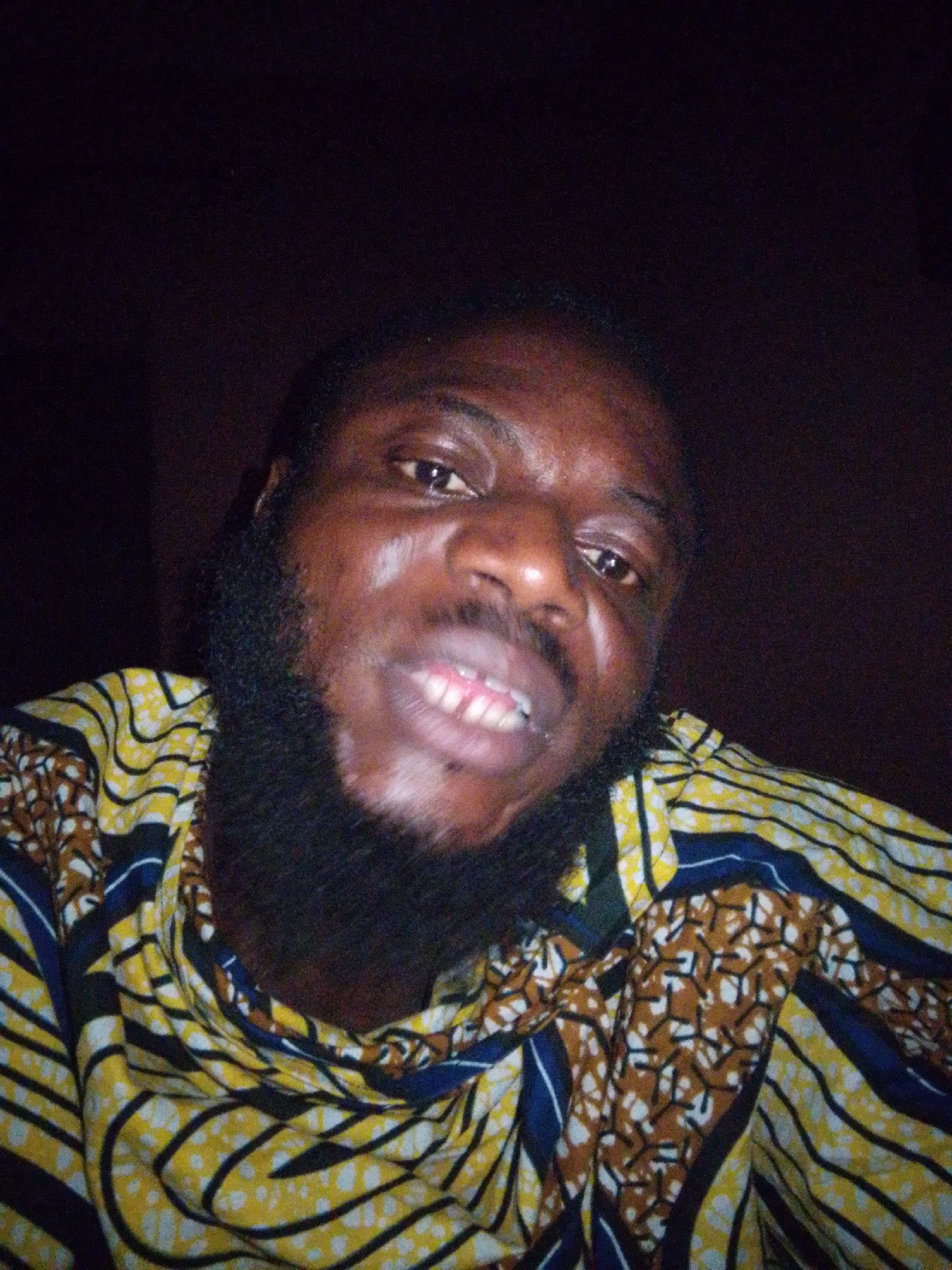 A close-up photo shows a man with dark skin, a full black beard, and short hair. He is wearing a patterned shirt with yellow, brown, and blue colors. He is looking at the camera and slightly smiling, revealing his teeth. The background is dark, suggesting an indoor setting with low lighting. There is no explicit information to determine if the photo was taken in Lokoja, Nigeria.
