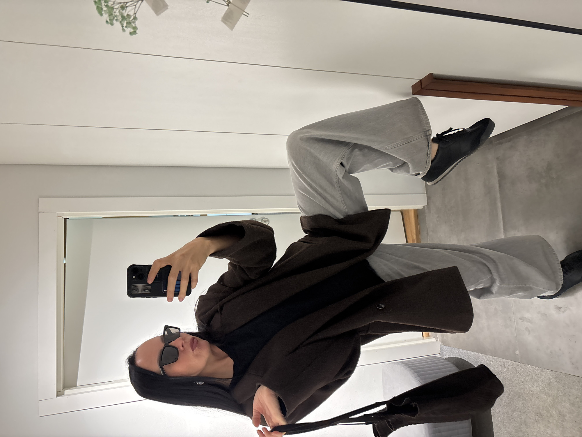 A woman in sunglasses stands in an indoor space, taking a selfie in a mirror. She wears a dark, buttoned coat, gray pants, and black shoes. She holds a phone in her right hand and a dark purse with straps in her left. The background includes a white wall with a plant on top, a door frame, and a light gray tiled floor. The location of the scene is not identifiable from the context of the image.