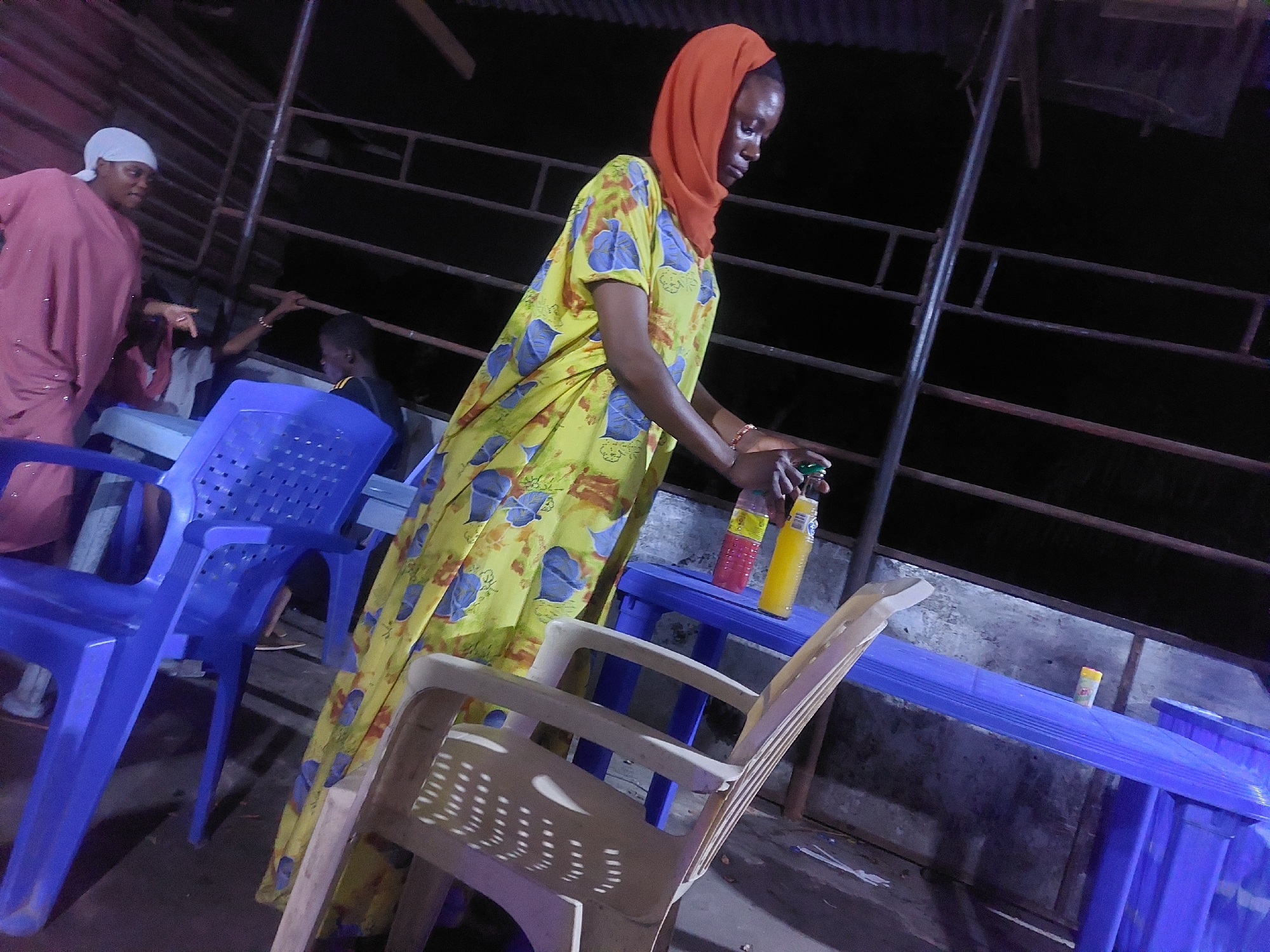 The image shows an outdoor scene in Dar es-Salaam, Tanzania, possibly a restaurant or food stall at night. A woman wearing a yellow and blue patterned dress and an orange hijab is placing bottles on a blue plastic table. Two bottles, one pink and one yellow, are visible on the table. Nearby, there is a blue plastic chair and a white plastic chair. In the background, another woman wearing a pink dress and a white head covering is gesturing towards a person sitting at a table. The setting appears to be under a corrugated roof structure with metal railings.
