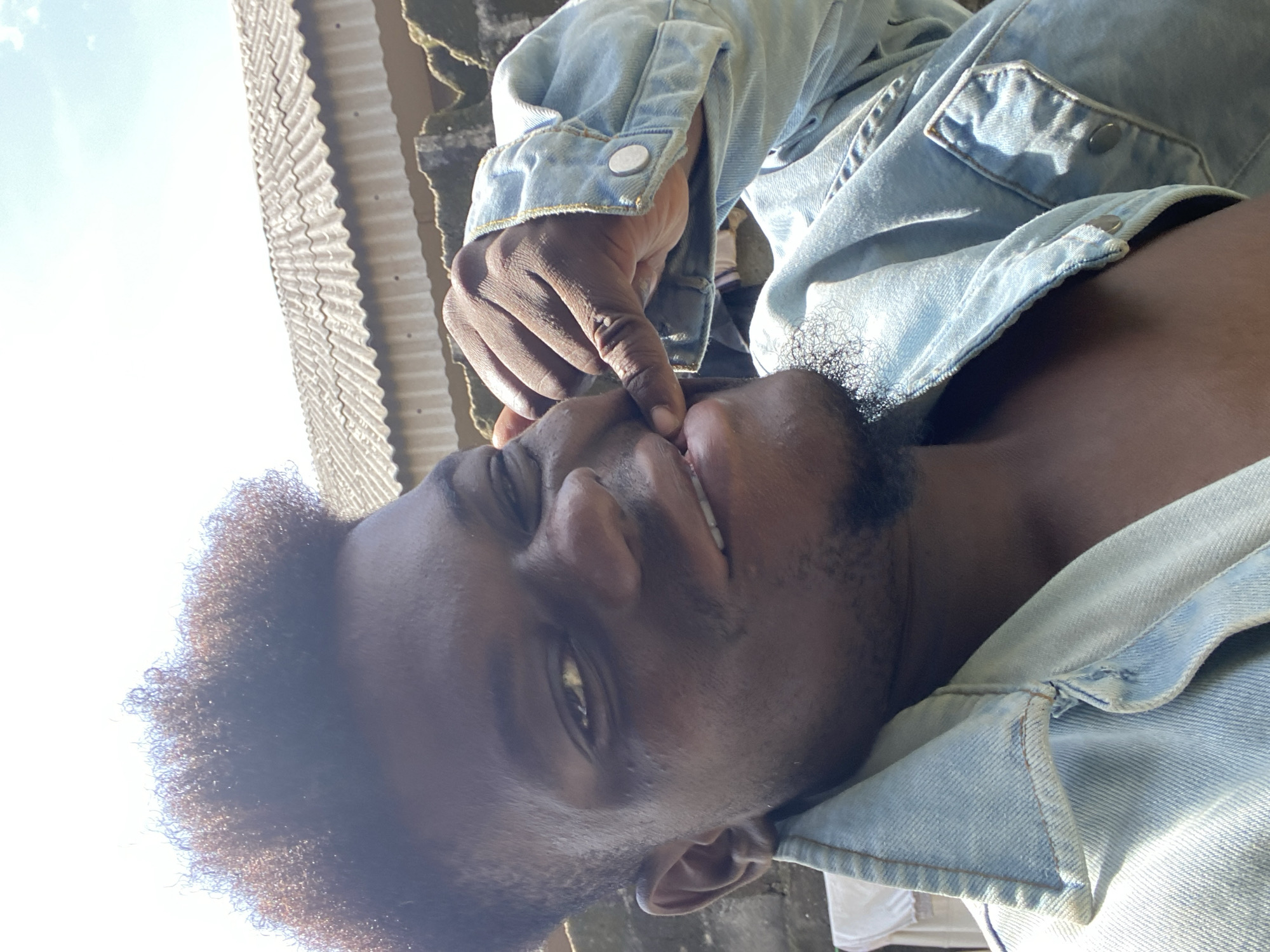 A man with dark skin is outside in Unknown, Unknown. He has dark, curly hair and a short beard. He is wearing a light blue denim jacket. He is holding a finger to his mouth and smiling slightly. The background includes a corrugated metal roof and a wall. The sky is visible in the background.