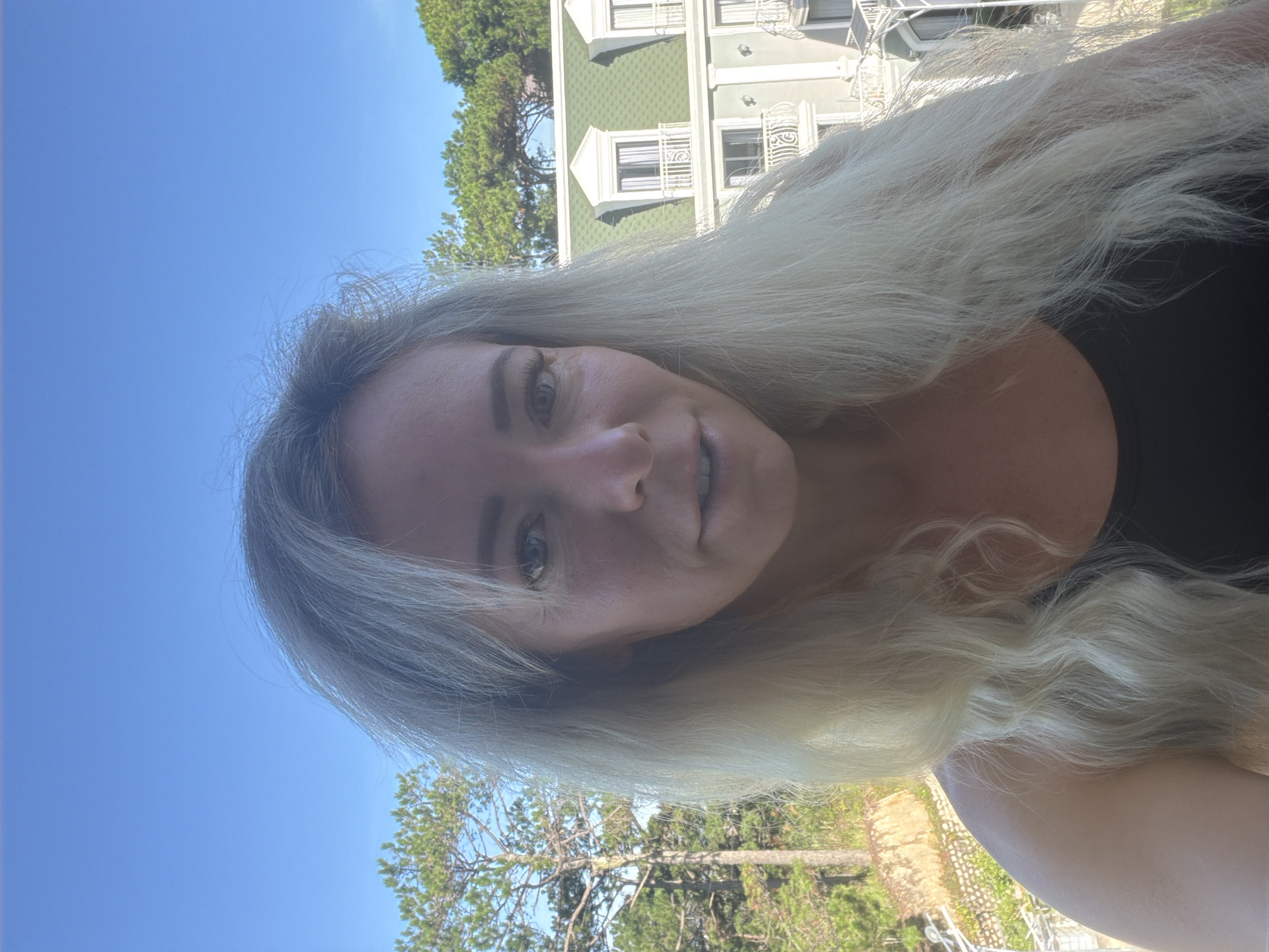 A woman with long blonde hair is taking a selfie outdoors. She is wearing a black tank top. Behind her, a building with a green roof and white trim is visible, along with trees and a clear blue sky. The setting appears to be a hillside with a view of architecture and nature.