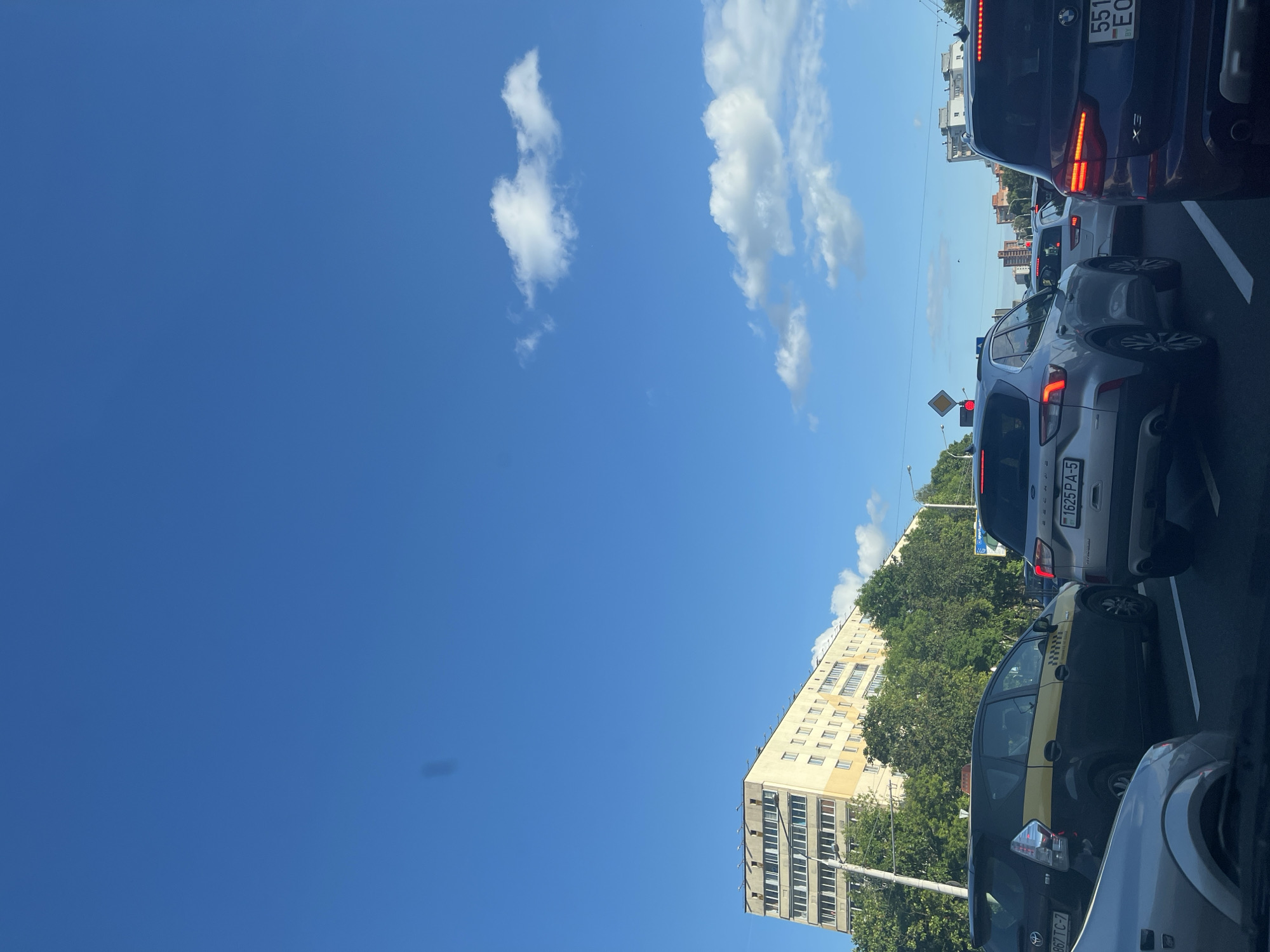 The image captures a sunny day in Minsk, Belarus, likely during the daytime, as indicated by the clear blue sky and bright sunlight. The scene depicts a traffic jam on a city street, with several cars lined up. The perspective is from inside a vehicle, looking forward and slightly upwards.
In the foreground, the back of a silver SUV is visible, with its license plate reading "1625 PA-5". Next to it, a black and yellow taxi is also stopped in traffic. Further back, other vehicles, including a dark blue BMW with a license plate displaying "551 EO", are visible in the congestion. The buildings lining the street include a tall, light-colored apartment building with many windows, characteristic of Soviet-era architecture. Lush green trees are interspersed between the buildings and along the roadside, suggesting a pleasant, possibly summer season. White clouds dot the bright blue sky.
The overall mood is one of a typical urban commute. The traffic suggests a moment of standstill, with drivers likely waiting for the congestion to clear. There are no people clearly visible, focusing the scene on the vehicles and the urban environment. The presence of the license plates with Cyrillic characters and the architectural style of the buildings strongly suggest the location as Minsk, Belarus.