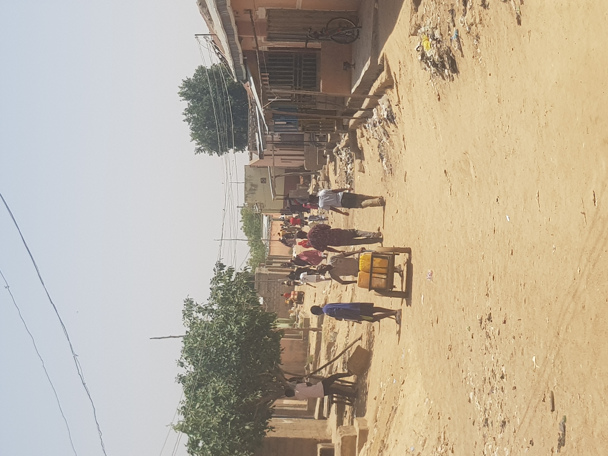 The image depicts a street scene in Dutsinma B, Nigeria. The road is unpaved, appearing to be composed of compacted dirt. Several people are present. One man is pulling a cart with yellow water jugs on it, with another man walking alongside. In the background, a group of people are gathered, possibly at a market or meeting point. Buildings line the road, constructed from what appears to be brick and plaster, with corrugated metal roofing. Overhead, power lines are visible. There is litter scattered along the side of the road. A tree is present in the foreground. The overall impression is a scene of daily life in a developing urban environment.