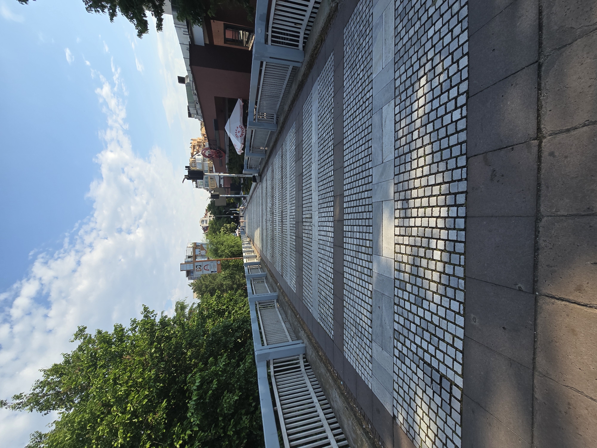 The image displays a bright, sunny outdoor scene, likely a wide sidewalk or promenade next to a lower area. The perspective is looking down the path, which is composed of alternating sections. On the right, large dark grey or brownish rectangular paving slabs form one part of the path. To their left, there's a section of smaller, light-colored square paving stones that resemble cobblestones. These two patterns repeat further into the distance.
A light grey railing or fence runs along the left edge of the paved path, separating it from a lower tier of dense green trees and foliage. Beyond the trees, various multi-story buildings, appearing to be residential or commercial, line the street. The sky above is a clear, bright blue with scattered white cumulus clouds.
The strong sunlight indicates it is likely midday or early afternoon, casting distinct shadows on the paving stones. No people or specific activities are visible, contributing to a calm atmosphere.
Several textual elements are present:
- A white flag or banner with red lettering, displaying "STELLA" and partially visible text below it, hangs from a building in the mid-ground.
- A yellow and white sign near the trees on the left clearly shows "B&C", with additional, smaller text underneath. Below this, another sign is partially visible with letters "M", "P", and what appears to be "MARKET".
- A red circular sign is visible on a building further down the path, though its text is not legible.