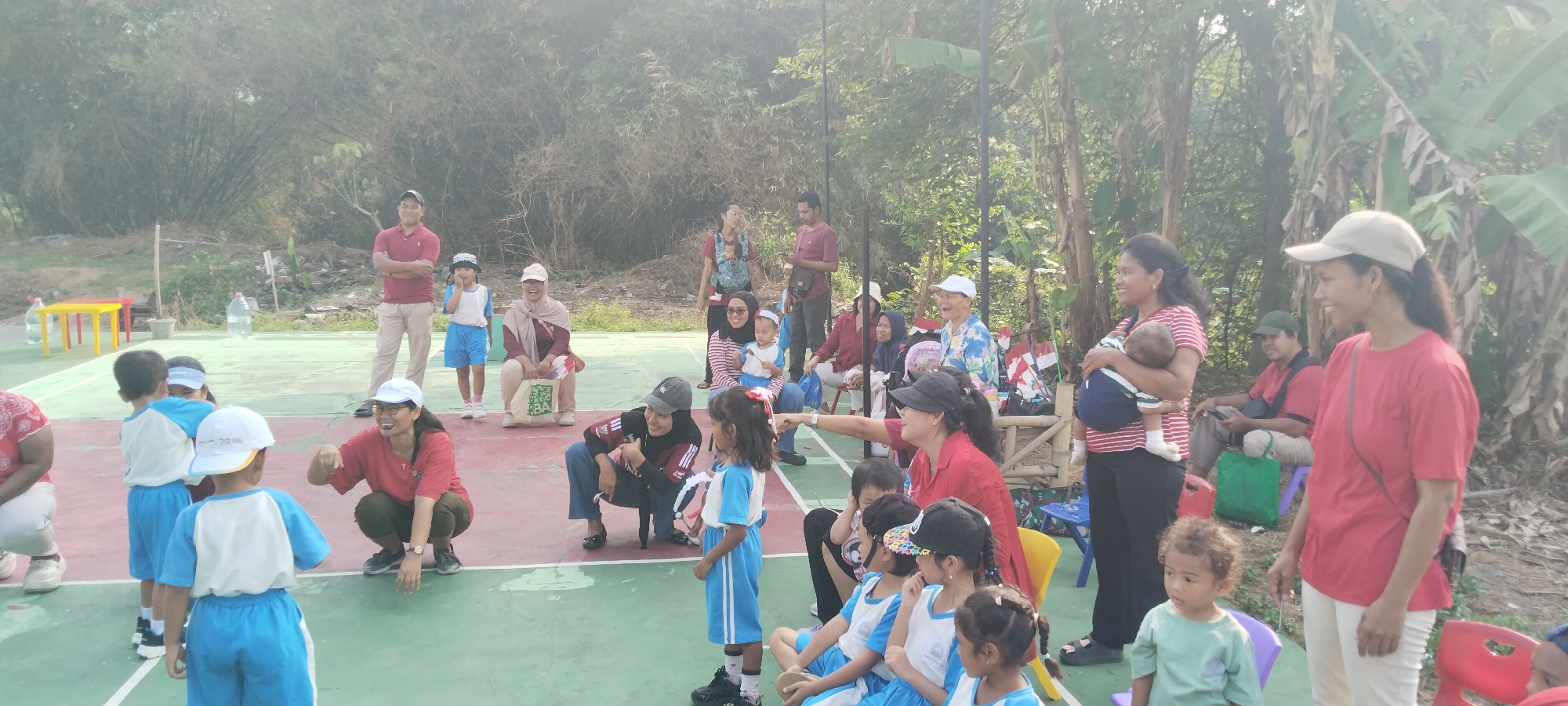 This image depicts an outdoor gathering, possibly a school event or sports day, taking place on a red and green court, likely a basketball court, in Taban, Indonesia. The scene is filled with a mix of adults and children, with many adults supervising or interacting with the children.
In the foreground, a woman in a red shirt and khaki pants stands looking to her left, wearing a light-colored baseball cap. To her left, a woman in a red top and dark pants is crouched down, gesturing towards a group of children. Several children, dressed in blue and white uniforms, are either standing or sitting on the court. One young child, a boy in a light green t-shirt, stands in the middle ground to the right, looking slightly upwards.
Further back, several adults are seated on chairs or benches. A woman in a red shirt holds a baby wrapped in a blue and white striped sling. Another woman, wearing a headscarf and a maroon outfit, is seated on the ground. A man in a red polo shirt and light-colored pants stands with his arms crossed, looking towards the children. Other individuals are visible in the background, including a woman in a blue shirt holding a baby, and a man in a maroon shirt.
The environment suggests a slightly humid climate, with lush green trees and vegetation visible behind the court. Bamboo stalks are present in the background to the left, and banana trees are prominent on the right. The lighting indicates it is daytime, possibly late afternoon, with the sun casting a warm glow on the scene. Scattered on the court are a few red and yellow tables, water bottles, and what appear to be small flags.