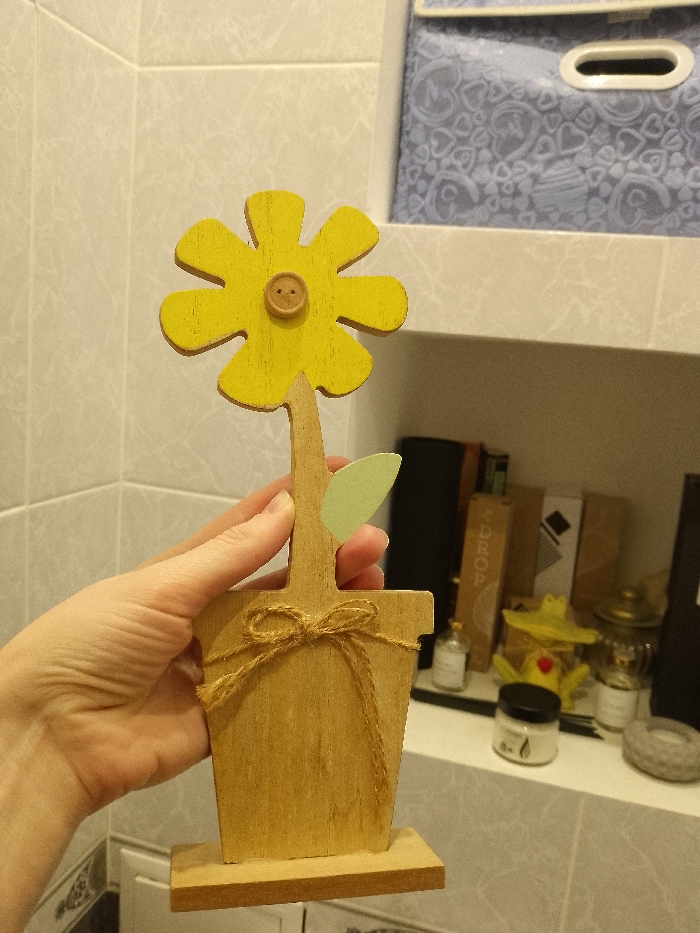 A light-skinned human left hand holds a decorative wooden flower against a background of light-colored wall tiles and white shelving. The wooden flower features a bright yellow, six-petaled head with a small, round, light brown wooden button as its center. The stem and flowerpot-shaped base are unpainted wood. A single light green wooden leaf is attached to the stem. Light brown twine is wrapped around the wooden pot and tied in a bow on the front. The object is held upright, slightly angled towards the viewer.
The background comprises rectangular light-colored wall tiles. Two white shelves are visible. On the upper shelf, a light blue soft-sided storage box with a white heart pattern and a white oval handle is present. The lower shelf displays several items: a small clear glass bottle, stacked brown and black objects (possibly books or decorative panels, one partially revealing "DROP"), a yellow ceramic object with a red detail, another small clear bottle, a black jar with a white lid, and a circular grey textured object. The setting is an indoor environment in the city of Unknown, Unknown.