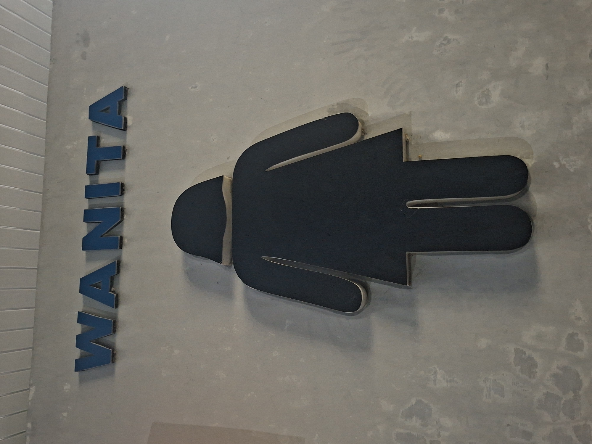 The image shows a sign for a women's restroom. The sign is blue and has the word "WANITA" which is Indonesian for "WOMEN" spelled out in capital letters above a black silhouette of a woman. The sign is mounted on a gray concrete wall. The wall has a textured finish and some visible stains and imperfections. Above the wall, there is a ceiling with horizontal white panels. The setting appears to be indoors, possibly in a public building or establishment. The city of Sinyeu, Indonesia is not explicitly mentioned in the image, but the word "WANITA" suggests it is located in Indonesia.