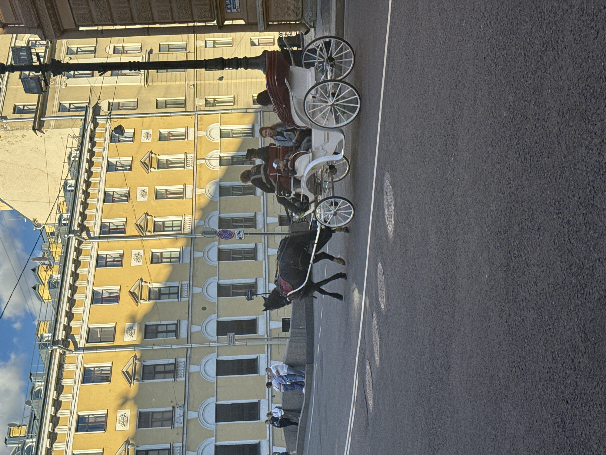 The image depicts a horse-drawn carriage on a street in Saint Petersburg, Russia. The carriage is white with red upholstery and is pulled by a dark-colored horse. Several people are riding in the carriage, including two adults in the back and one child and one adult in the front. In the background, there is a yellow building with multiple windows and architectural details. The street is paved with asphalt, and there are manhole covers visible on the road. Several people are standing on the sidewalk in the background, observing the scene. The sky is blue with some clouds, and the sunlight casts shadows on the buildings. The presence of the horse-drawn carriage and the architecture suggest a historical or tourist area within the city.