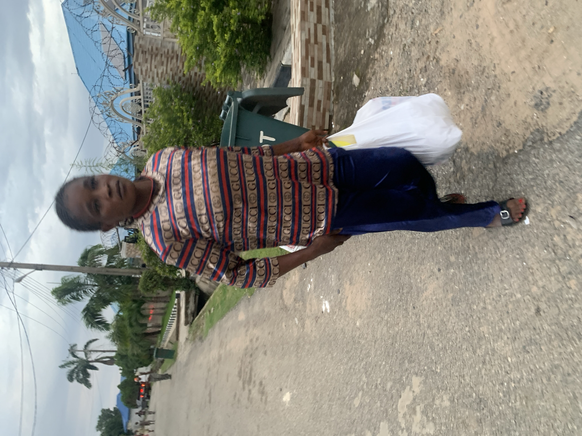 A full-body, medium shot captures a Black woman walking on a paved road in Abuja, Nigeria. She is wearing a long-sleeved, multi-colored striped shirt with a repeating "GUCCI" logo pattern, dark blue pants, and black sandals. She is carrying a large white plastic bag in her right hand. The background shows a street with a few palm trees, buildings with blue roofs, and a green trash bin. The sky is overcast.