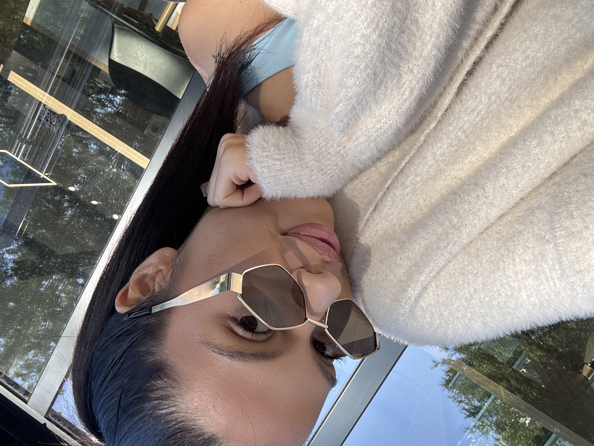 A close-up selfie of a woman with dark hair pulled back in a ponytail, wearing gold-rimmed sunglasses and a fluffy cream-colored sweater. She is leaning her head on her hand, with her lips slightly parted. The background shows a reflection of trees and sunlight on a window, suggesting an outdoor or well-lit indoor setting. The image is framed by the reflection of a modern building with wooden accents and black chairs. The woman has a small tattoo on her shoulder. The scene is likely outdoors, possibly in a cafe or a public space with large windows. The overall lighting suggests it's daytime. No specific location in Sofia, Bulgaria, can be identified from the visual information.