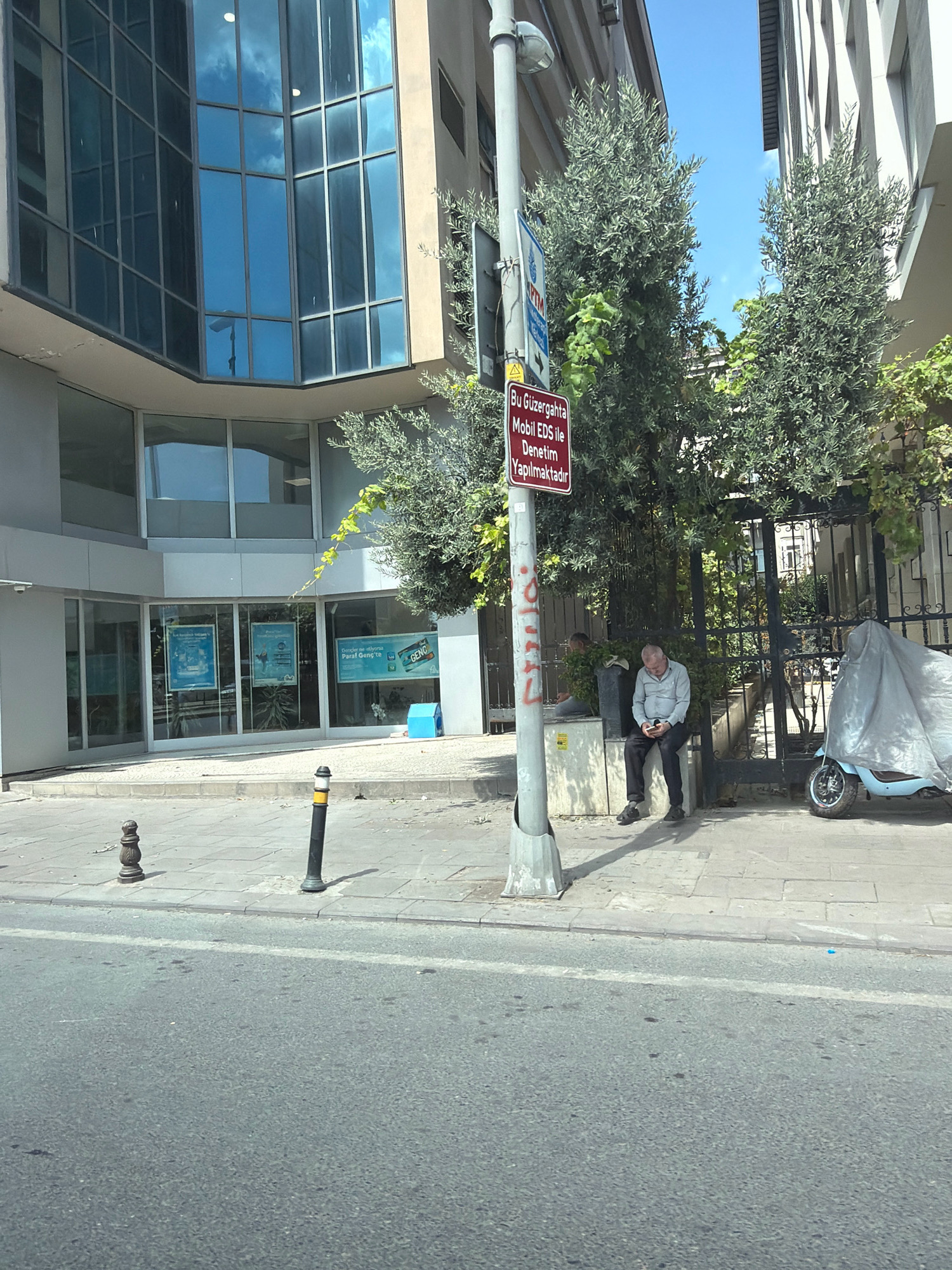 A man sits on a concrete ledge next to a street in Bahçelievler, Turkey, looking at his phone. He is wearing a light-colored long-sleeved shirt and dark pants. Beside him is a light blue scooter covered with a light gray tarp. Behind the man is a dark metal gate and trees. A red sign is mounted on a pole in front of the trees. The street in the foreground is asphalt with a white line. The sidewalk is made of gray paving stones. To the left of the sidewalk is a building with large glass windows.