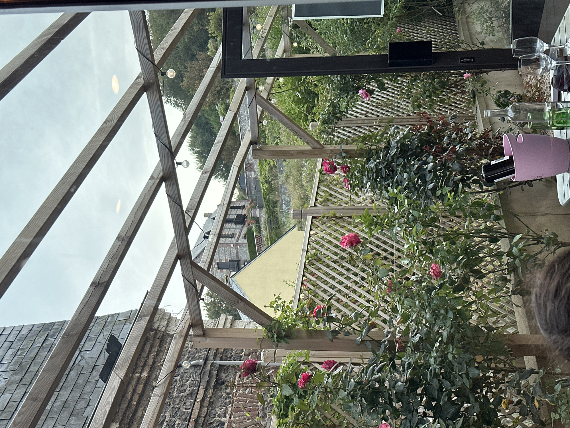 This image captures a view from a covered outdoor seating area, possibly a restaurant or cafe, in Veules-les-Roses, France. The scene is characterized by a wooden pergola structure overhead, with exposed beams and string lights. In the foreground, several rose bushes with pink blooms are visible, intertwined with a lattice fence. A table is set with wine glasses and bottles, including an ice bucket containing bottles. Behind the roses, a stone building with a slate roof and a yellow house are partially visible on a hillside, suggesting a village setting. The overall atmosphere is that of a charming, possibly tranquil, outdoor dining space with a hint of natural beauty.