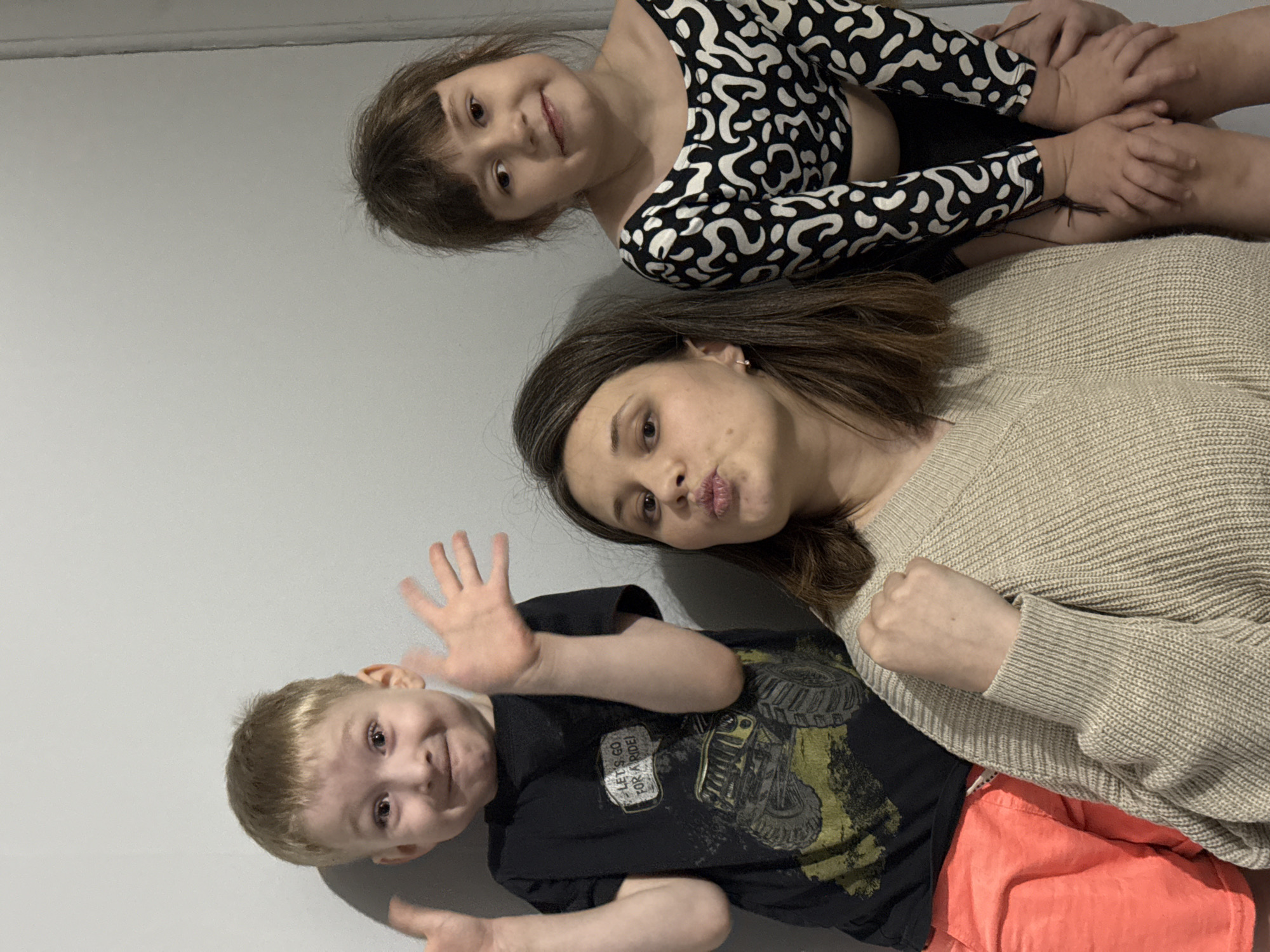 The image captures a playful indoor family moment in Chaikovsky, Russia, featuring an adult woman and two young children, a girl and a boy. The plain light-colored wall behind them serves as a neutral backdrop.
The adult woman, positioned centrally, has brown hair and wears a light beige knitted sweater. She is looking towards the camera with her lips puckered playfully. Her right hand is lightly clenched in a fist.
To the woman's left, a young boy with light hair is making a goofy face, his eyes wide and mouth slightly open in a mischievous smile. Both of his hands are raised with palms open. He is wearing a dark t-shirt with a graphic of a monster truck, featuring the text "LET'S GO FOR A RIDE!", and red/orange shorts.
Above and slightly to the right of the woman, a young girl with brown hair and bangs is smiling at the camera. She is dressed in a black and white abstract patterned long-sleeved top. Her hands are clasped together near her knees, suggesting she is seated or kneeling.
The overall mood of the photograph is lighthearted and joyful, capturing candid, silly expressions from the family members. The lighting suggests an indoor environment.