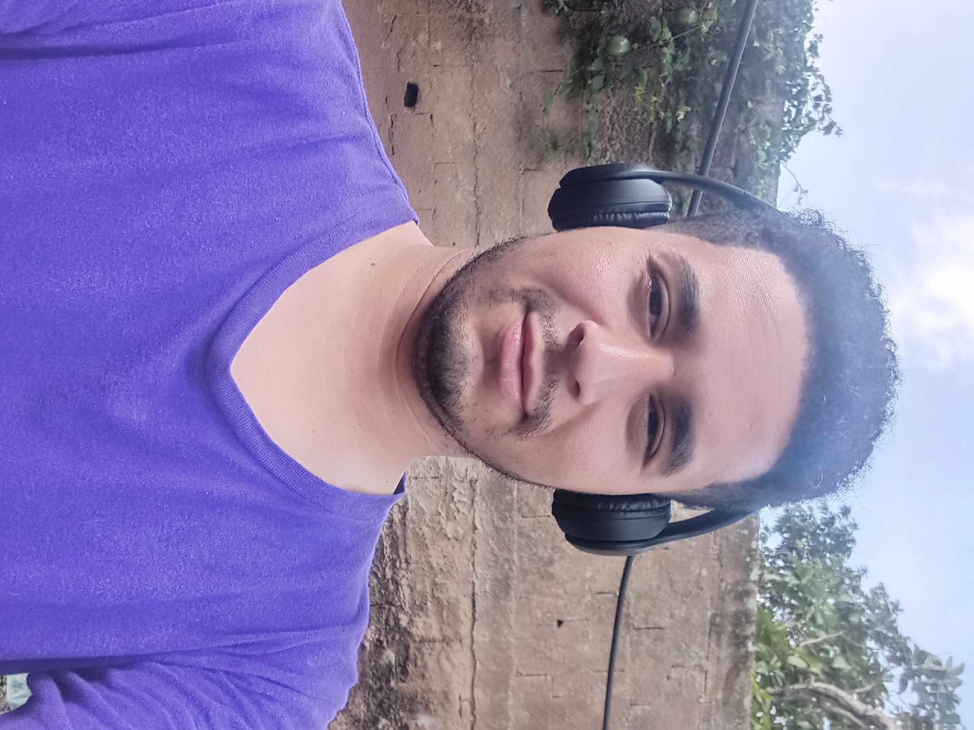 In Duaca, Venezuela, a man with dark, curly hair and light brown skin is shown in a selfie. He is wearing black headphones and a vibrant purple t-shirt. The background consists of a textured, light brown brick wall and some greenery. The sky above is a pale blue.