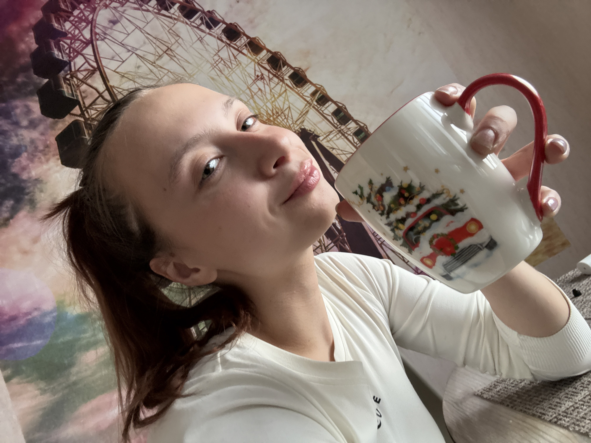 A young woman with light skin, brown hair pulled back in a ponytail, and slightly glossy lips is holding a white mug with a red handle and a Christmas-themed design in her right hand, appearing to drink from it. She is wearing a long-sleeved white shirt. The background features a wallpaper depicting a Ferris wheel and what appear to be sky elements. There are no identifiable details indicating that the location is specifically in the city of Прикамский, Russia, beyond general indications of an indoor domestic environment.