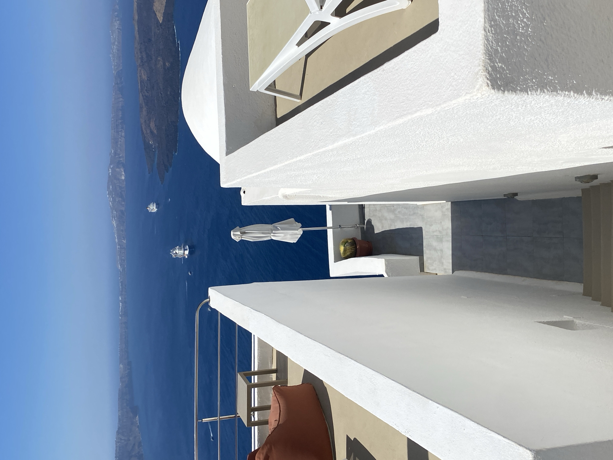 A high-angle, full shot captures a bright, sunny day overlooking the Aegean Sea from Thira, Greece. The foreground is dominated by the whitewashed architecture characteristic of the Cycladic islands, with a sharp white wall dividing the frame. To the left, a section of a patio is visible, featuring an orange beanbag chair and a small grey side table. Further right, a pathway leads down between two white walls, with a closed white umbrella and a potted cactus visible. Across the sea, the caldera of Santorini is apparent, with a large cruise ship and other smaller vessels navigating the deep blue water under a clear sky. The image conveys a sense of leisure and breathtaking natural beauty.