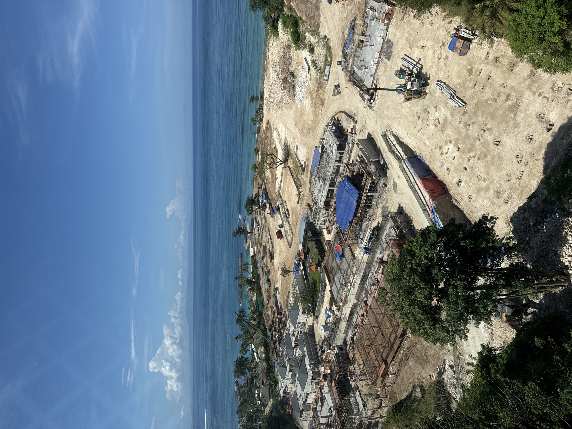 The image depicts a construction site along the coast in Kuta Selatan, Indonesia. Visible are partially constructed buildings with scaffolding and blue tarps, a crane, and construction equipment. A container is also visible. The scene includes trees, dirt roads, and the ocean. The sky is blue with some clouds. The construction appears to be taking place near the coastline.