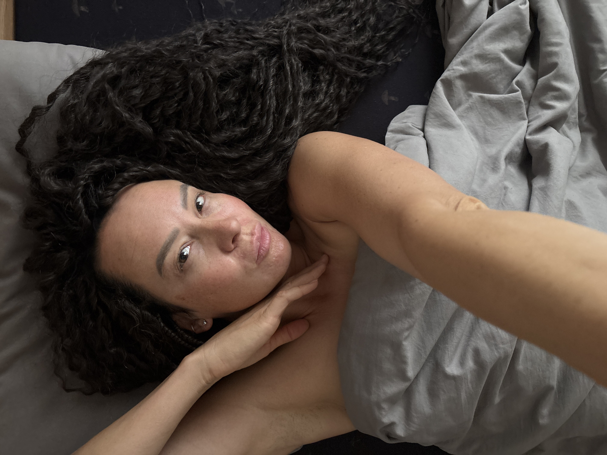 A woman with long, dark, curly hair is lying in bed under a gray duvet. She has fair skin, freckles, and is wearing a small earring. Her right hand is touching her neck and the other arm is extended in what appears to be taking a selfie. The setting is presumably indoors, in a bedroom. There is no indication of the city of Chaikovsky, Russia, in the image.