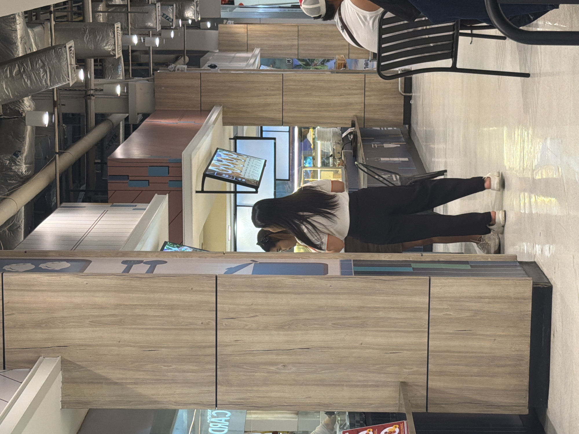 The image depicts an indoor scene, likely a food court or shopping mall. Two women stand near a counter, with one looking towards a digital menu board. The environment features wood-paneled structures, polished floors, and overhead ductwork. In the background, a person wearing a white shirt and a red and white cap is seated at a black chair. The scene appears to be from a public space in Ban Rang Rak, Thailand, given the context of the request.