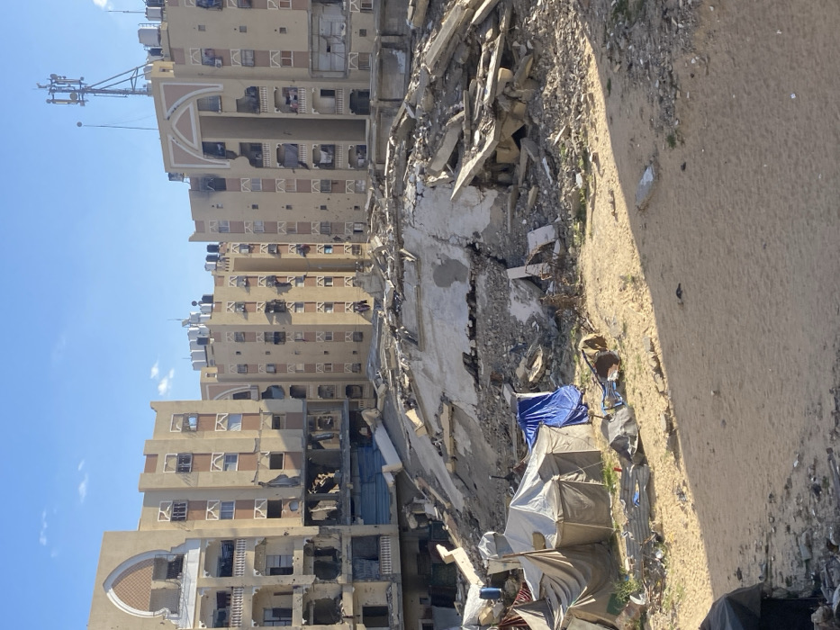 The image depicts a scene of extensive urban destruction in Khan Yunis, Palestinian Territory, under a clear blue sky. In the background, several multi-story residential buildings, primarily light brown or tan with terracotta-colored accents, exhibit significant damage. These structures feature numerous broken windows, facades heavily pockmarked with impact holes, and visible structural damage including collapsed sections and exposed interiors. Some balconies still have laundry hanging, and black water tanks are visible on rooftops, along with a communication tower on one building.
The midground is dominated by a large, irregular mound of pulverized concrete, rebar, and general construction debris, indicating the complete collapse or severe destruction of a building. This rubble heap is extensive and covers a wide area.
In the foreground, a sandy, unpaved ground surface is present. To the left, a small, makeshift encampment consists of several tents constructed from various tarpaulins and fabrics, including one distinct blue tarp. Scattered debris, such as corrugated metal sheets, a blue plastic barrel, and what appears to be discarded furniture, is visible near the tents and across the sandy ground. No individuals are discernible in the frame, and there are no direct actions or interactions captured. The scene is illuminated by natural daylight, casting shadows.