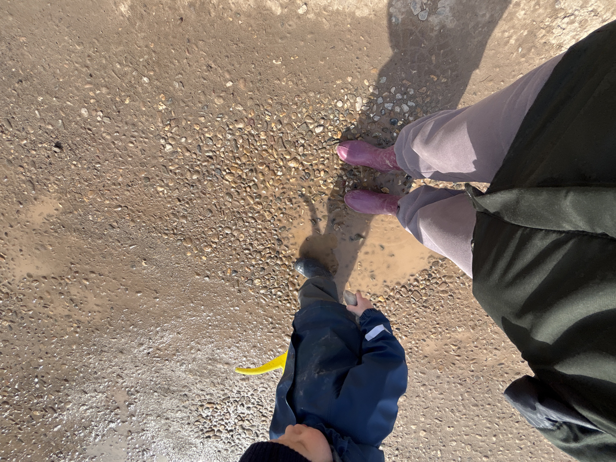 The image shows a top-down view of three people standing on a muddy, pebbled ground in an outdoor environment. One person is wearing pink rubber boots with light gray pants and a long dark green coat. A child is wearing a dark blue waterproof suit with a yellow plastic shovel. The third person has a dark sweater. The location cannot be determined from the image, and there is no verifiable information about the city of Ферма, Russia.
