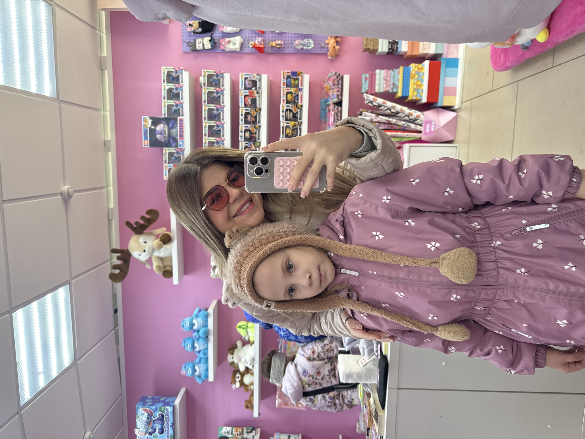 The image captures an indoor scene within a retail store in Chaikovsky, Russia, characterized by bright pink walls and white ceiling tiles with fluorescent lighting. In the foreground, an adult female with long light brown hair, wearing red-tinted sunglasses and a light-colored jacket, is on the right, smiling and holding a grey smartphone with a pink circular grip in her right hand, actively taking a selfie. Her left hand rests on the shoulder of a young girl, positioned center-left. The girl, with light brown hair and blue eyes, wears a pink hooded jacket with white floral patterns and a light brown knitted hat with long ear flaps ending in fuzzy pom-poms; she looks slightly off-camera to her right with a neutral expression. The background features white floating shelves displaying a variety of merchandise, including plush toys such as a moose, blue monster figures, and dog-shaped plushies, alongside numerous boxed Funko Pop! figures, prominently featuring characters like Stitch. A white counter on the left holds assorted small items, and further back, another individual wearing a floral jacket and grey headband is visible, facing away from the camera. A purple pegboard display with small accessories and stacks of colorful gift boxes are visible on the right side of the frame.