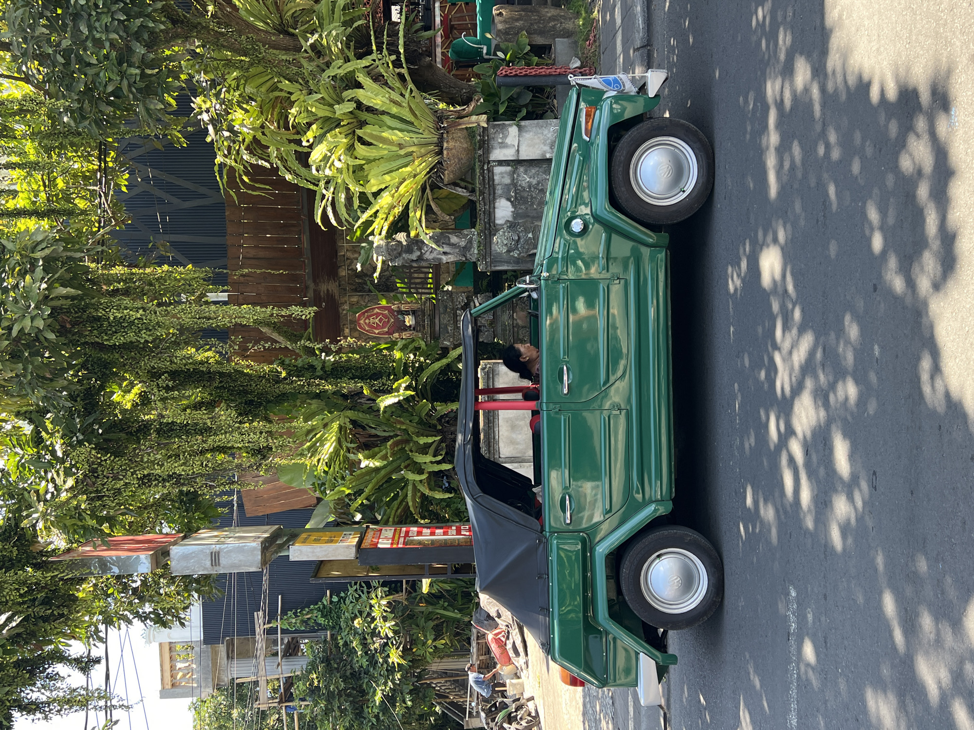 A vibrant green, open-top vehicle, resembling a Volkswagen Type 181 (The Thing), is parked on the side of an asphalt road in bright daylight. A person with dark hair is visible in the driver's seat, looking forward. The car has a black convertible soft top, currently folded down, and features silver-colored hubcaps with a visible "VW" (Volkswagen) logo on the front wheel. A small blue and white flag or decoration is attached to the front right side of the vehicle.
The surroundings are lush and tropical, dominated by dense green foliage, including large ferns and various trees. Dappled shadows from overhead trees fall distinctly across the sunny road surface, indicating a clear, sunny day, likely during the morning or late afternoon.
On the left, behind the vehicle and through the trees, a building with horizontal wooden slats and corrugated metal siding is visible. In front of this, traditional Balinese-style stone structures and carvings, along with a large bird's nest fern, contribute to the local aesthetic. A decorative red and white emblem is mounted on a stone pillar near the building. Further back and to the left, a person on a scooter or motorcycle is visible, along with what appears to be a red gas canister or tank on the ground. Various signs, including some with Indonesian script (e.g., "MIE AYAM...", "WARUNG..."), are partially visible but largely illegible in the background, hinting at local businesses. The scene strongly suggests a setting in a tropical, culturally rich area, consistent with the Ubud District in Indonesia.