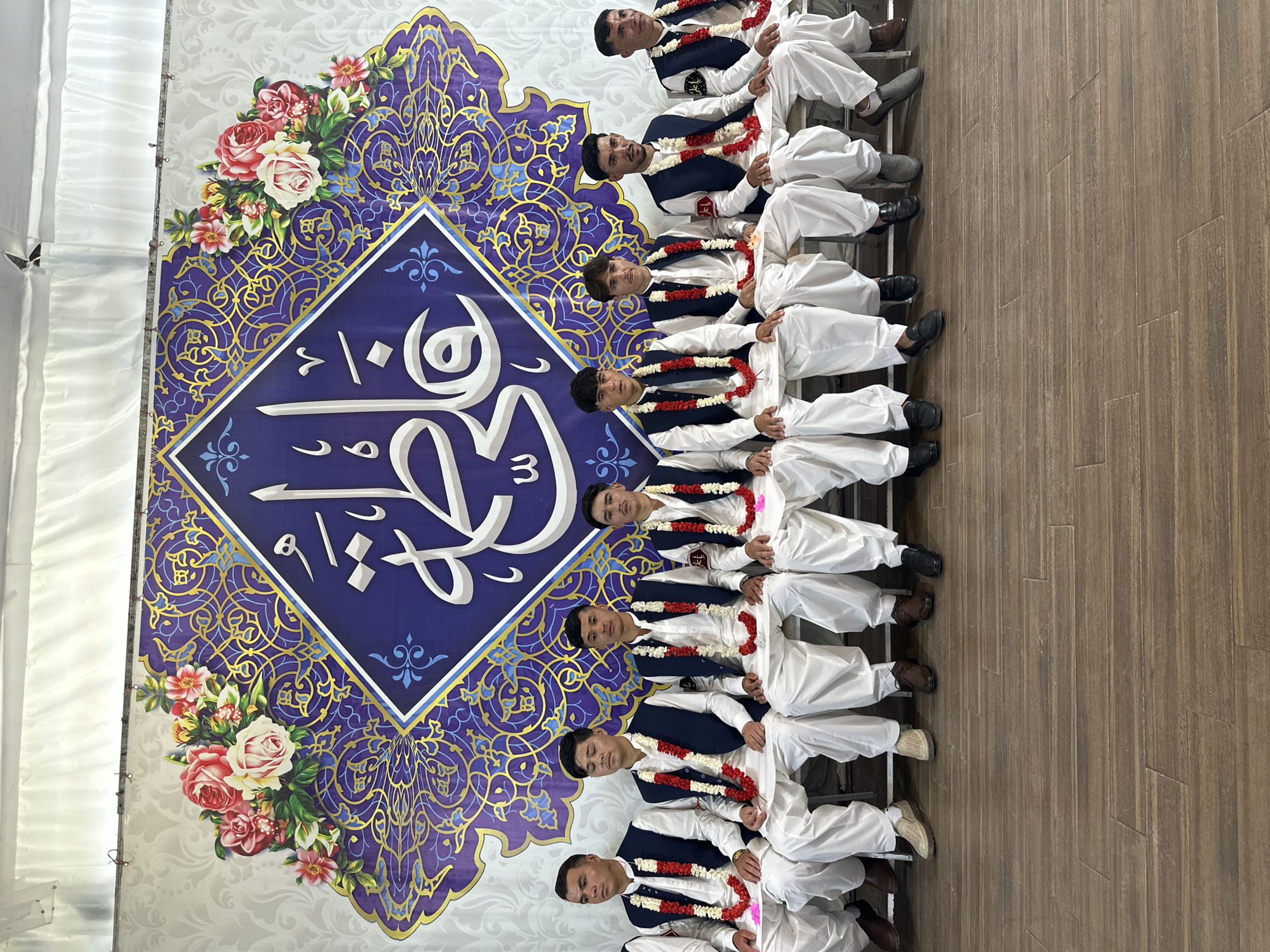 A group of nine males, likely young adults, are seated on steps in front of a backdrop featuring Arabic calligraphy and floral designs, possibly in Quetta City Tehsil, Pakistan. They are wearing matching traditional attire consisting of white shalwar kameez and dark blue vests, each adorned with a red and white floral garland. The backdrop is a mix of white, blue, and gold colors. The steps appear to be made of a wood-like material.