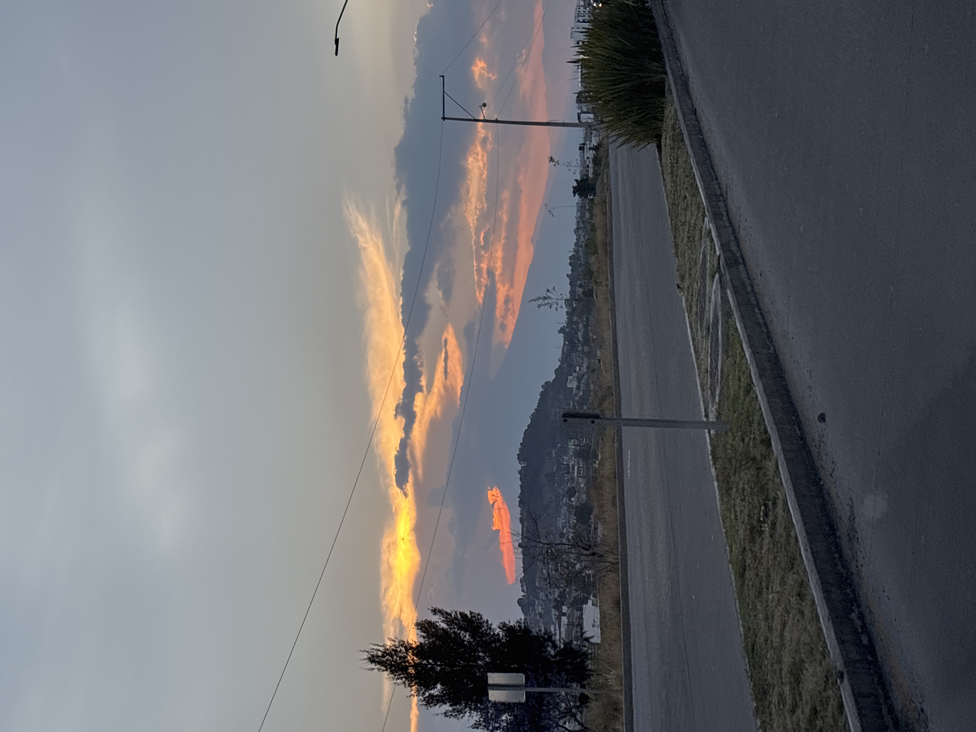 A landscape captures a serene sunset over Santa Clara Ocoyucan, Mexico. The foreground features an asphalt road with a grassy shoulder and a concrete curb. A lone tree stands to the left of the road, silhouetted against the bright sky. Overhead power lines stretch across the sky. A distant mountain ridge is visible, with buildings nestled against its slopes. The sky is a dynamic mix of pale blue and dark grey clouds illuminated with streaks of orange and yellow from the setting sun. A tall, green bush stands to the right.