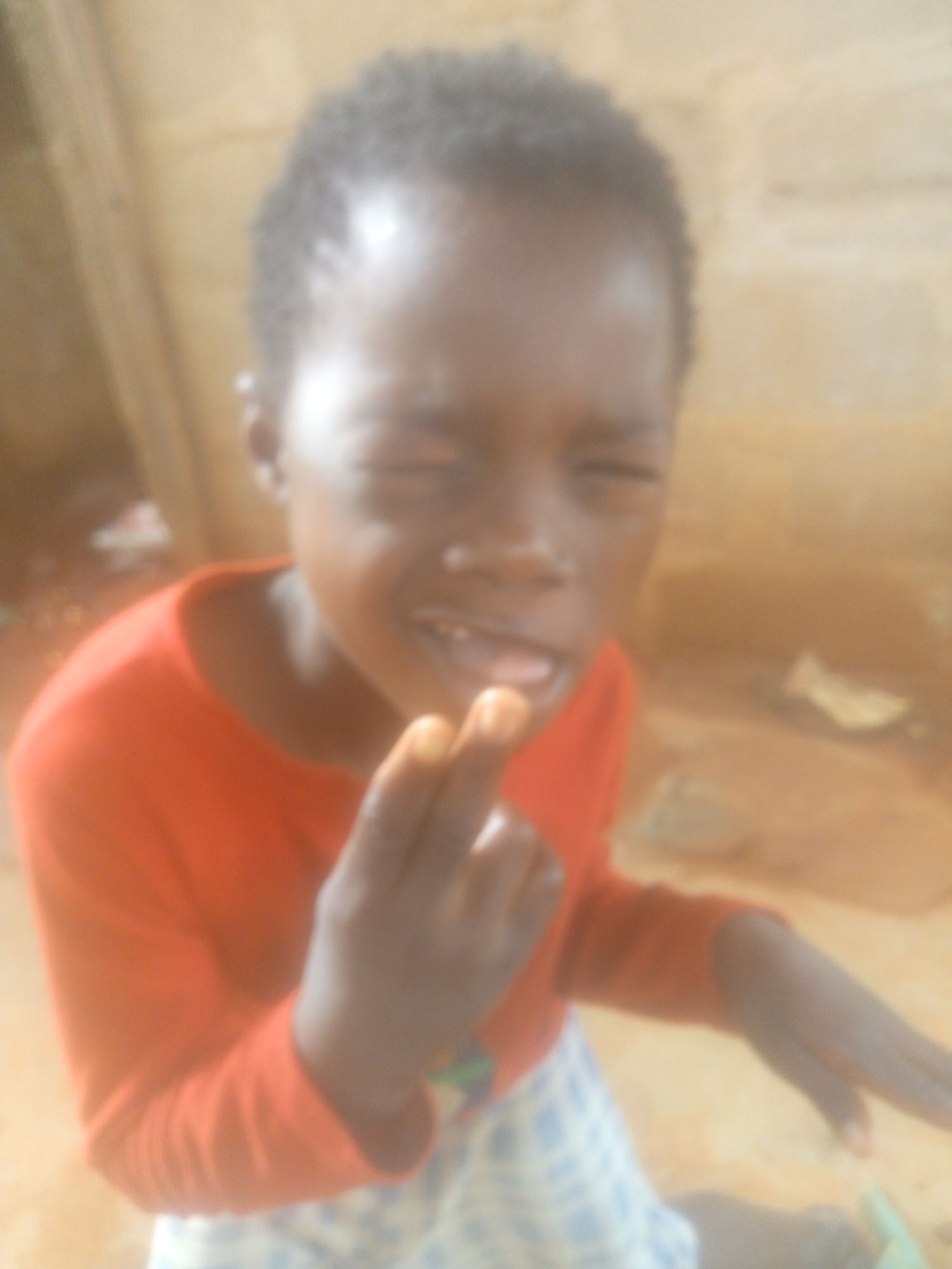 A slightly blurred, medium close-up shot features a young child, possibly a girl, with dark skin and short, dark, curly hair. The child is wearing a bright orange long-sleeved shirt and a blue and white patterned skirt. Their eyes are closed or tightly squinted, and their mouth is open, revealing some teeth and the tongue, suggesting a strong emotional expression. One hand is raised with fingers curled, positioned close to their mouth. Bright reflections are visible on the child's forehead and cheek, possibly indicating tears or sweat in the light. The child's other hand rests on the ground. The background is out of focus but appears to consist of earthen-toned walls or surfaces, implying an indoor or sheltered setting, possibly a simple dwelling. No text is visible in the image.