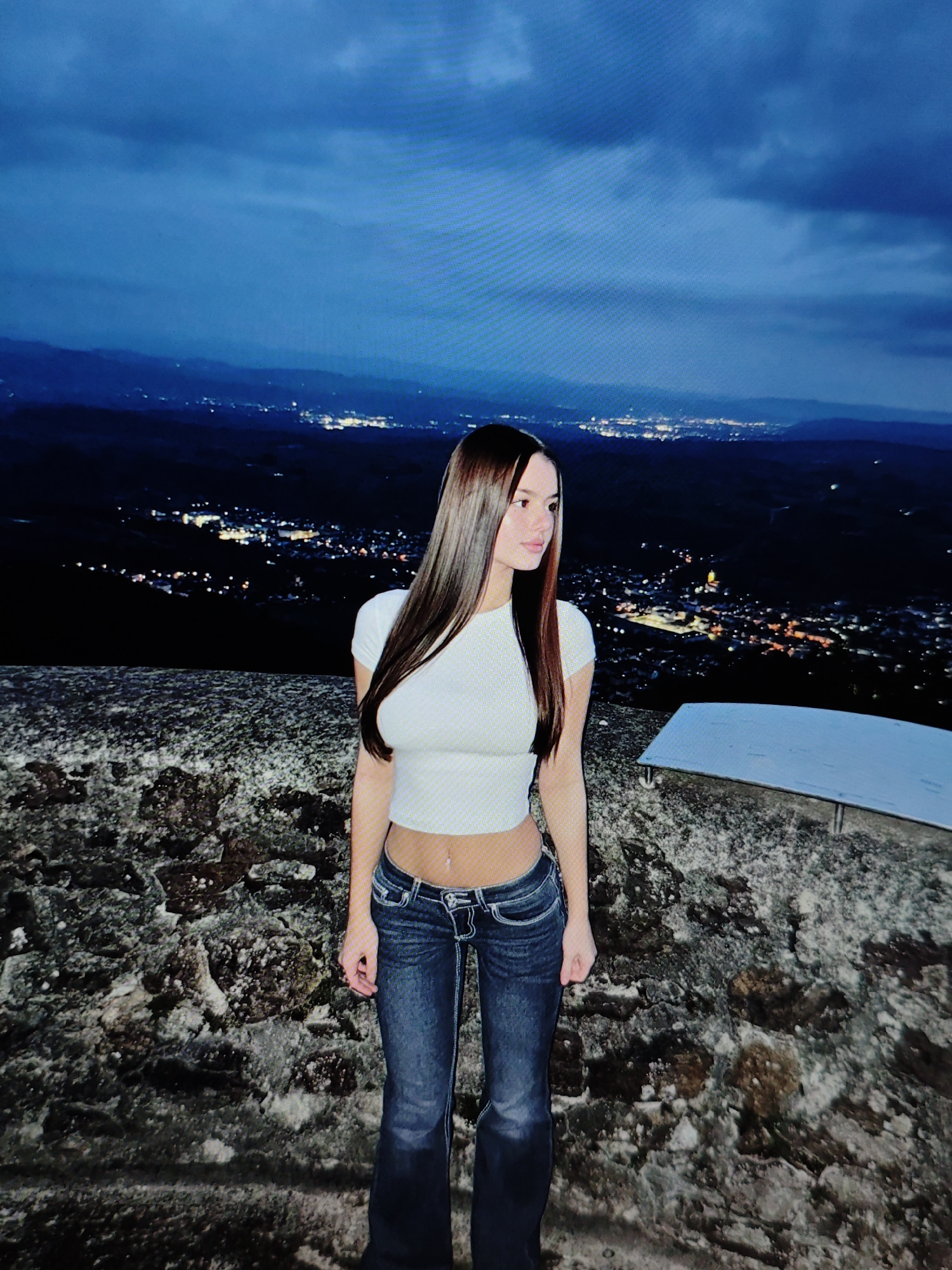 A young woman stands on a stone-laden surface, gazing out at a city illuminated at night. She is wearing a white crop top and blue jeans. The backdrop is a panoramic view of a city with numerous lights, suggesting it is a populated urban area. The sky above is dark blue with visible clouds, indicating it is nighttime. The overall scene conveys a sense of a serene evening observation point overlooking a vibrant city. The location is identified as Baku City, Azerbaijan.