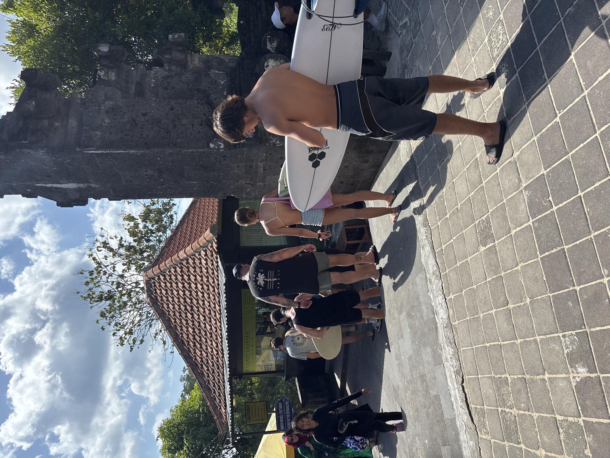 The image depicts an outdoor scene in bright daylight at what appears to be an entrance or gathering point in Kuta Selatan, Indonesia.
In the foreground, a young man with brown hair, shirtless, wearing dark grey board shorts and black flip-flops, holds a white "CHANNEL ISLANDS" surfboard under his right arm. He faces slightly left, casting a long shadow to his left. The ground consists of grey rectangular and square pavers with light grout lines.
To the left of the foreground man, a woman with her back to the camera, wearing a light pink halter top and light blue shorts, walks towards the mid-ground.
In the mid-ground, a group of approximately eight individuals is gathered near a ticket counter. The ticket counter is a wooden structure with a red tiled roof and a white corrugated awning. Visible signs on the counter include "TICKET COUNTER," "CAUTION!!!," and "MOHON MEMBELI TIKET MASUK" (Please buy entrance ticket) with an arrow pointing right. People are ascending steps towards this counter. Among them is a man in a black "SWELL" tank top and dark shorts, and another man in a light grey t-shirt carrying a circular, flat, light-colored object under his arm. On the far left, a young woman in black attire and pink shoes walks up a lower step, and behind her, a woman in a green patterned dress and a dark headscarf stands next to a partially visible yellow umbrella.
To the right of the mid-ground, a tall, dark grey, weathered stone pillar or gate structure stands prominently. Behind it and the buildings, lush green tropical foliage is visible.
The background features a bright blue sky with scattered white cumulus clouds. Sunlight is strong, casting distinct shadows of people and structures onto the paved ground.