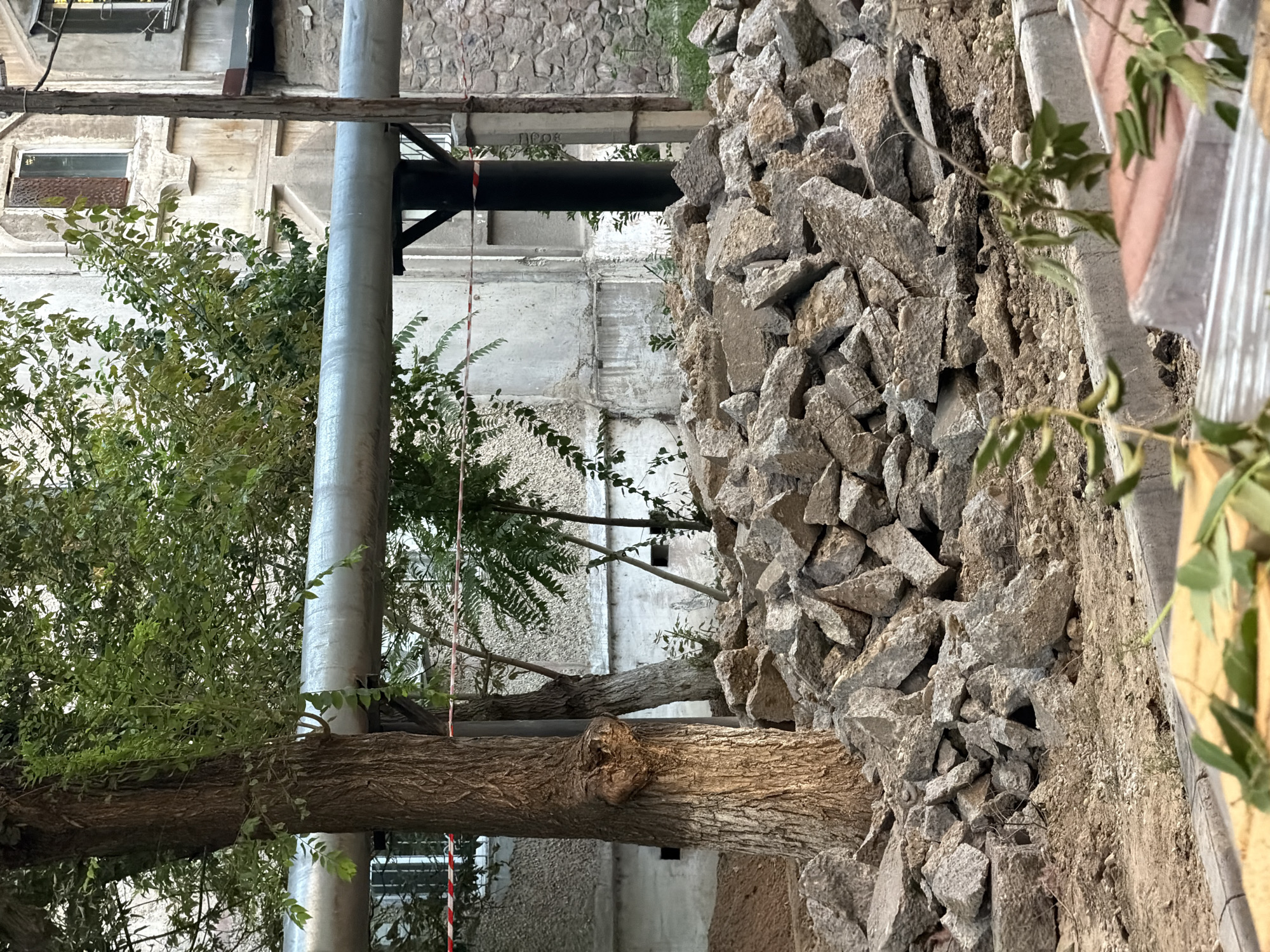 The image depicts an outdoor scene in Shymkent, Kazakhstan, characterized by construction debris and urban infrastructure. In the foreground, a large pile of broken concrete and rocks is visible, suggesting recent demolition or excavation work. A thick tree trunk stands to the left, its bark textured and weathered. Behind the debris and tree, a long, metallic pipe runs horizontally across the frame, supported by dark posts. The background features a weathered building with visible windows and rough stone walls, indicative of an older urban structure. A red and white striped tape runs across the scene, possibly marking a construction zone. Several smaller trees and green foliage are scattered throughout the image, indicating a natural element within the otherwise man-made environment. The lighting suggests it is daytime, with soft shadows present.