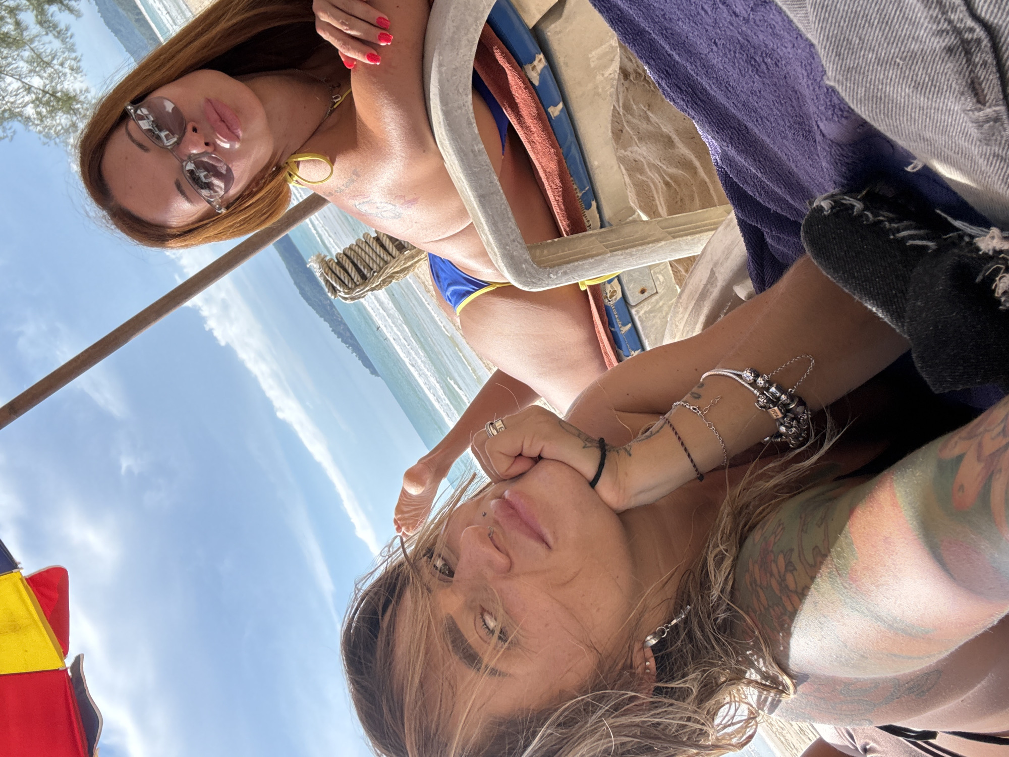 Two women are lying on beach chairs under a beach umbrella on a sandy beach in Choeng Thale, Thailand. The woman in the foreground is lying on her side, looking at the camera. She has blonde hair and is wearing a black bikini top. She has tattoos on her arms and chest. The woman in the background is lying on her back, looking up at the camera. She has long brown hair and is wearing a yellow bikini top and blue bikini bottoms. She is wearing sunglasses and has red nail polish on her fingernails. The ocean is visible in the background, with waves crashing on the shore. The sky is blue with some clouds. The beach umbrella is red, yellow, and blue.