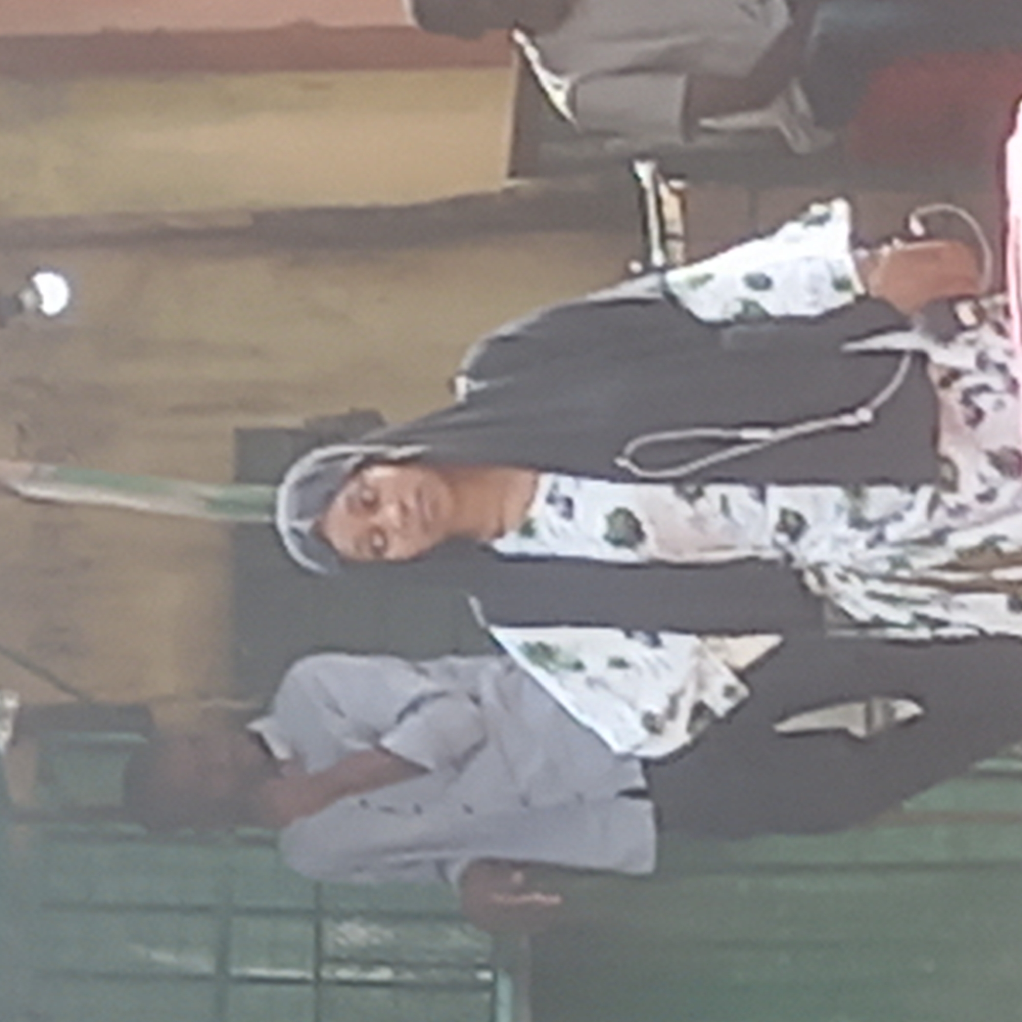 The image depicts a woman wearing a hijab and a floral-patterned dress with a black overlay. She is standing in an outdoor setting with a man in a grey shirt standing behind her. The location appears to be outdoors based on the visible backdrop. No specific structures or landmarks confirm the exact location, Dadi Riba, Nigeria. The woman is the central figure, and the man in the grey shirt is the secondary subject. The overall environment appears to be an outdoor gathering or public space.