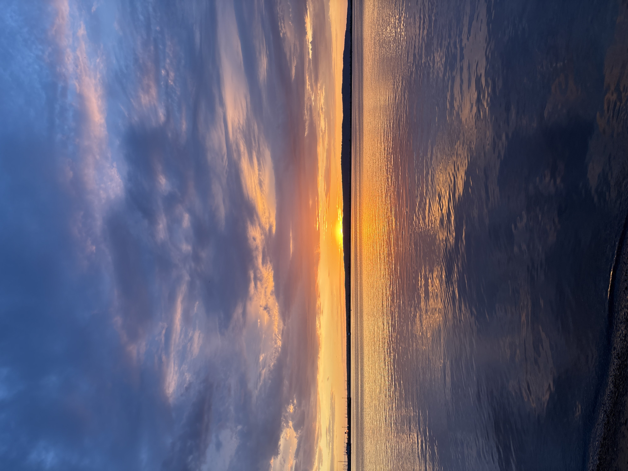 The image depicts a serene sunset over the Kama River in Chaikovsky, Russia. The sky is a dramatic canvas of deep blues, purples, and vibrant oranges, with clouds reflecting the warm hues of the setting sun. The sun itself is a bright, glowing orb positioned just above the horizon, casting a golden light across the water. The river's surface is calm, with gentle ripples that mirror the colors of the sky, creating a breathtaking reflection. The far bank of the river is visible as a dark, silhouetted line against the illuminated sky, with faint outlines of industrial structures and distant lights suggesting the presence of the city. The foreground shows a dark, sandy shoreline with a hint of water lapping at its edge. The overall impression is one of tranquility and natural beauty, capturing a picturesque moment in Chaikovsky.