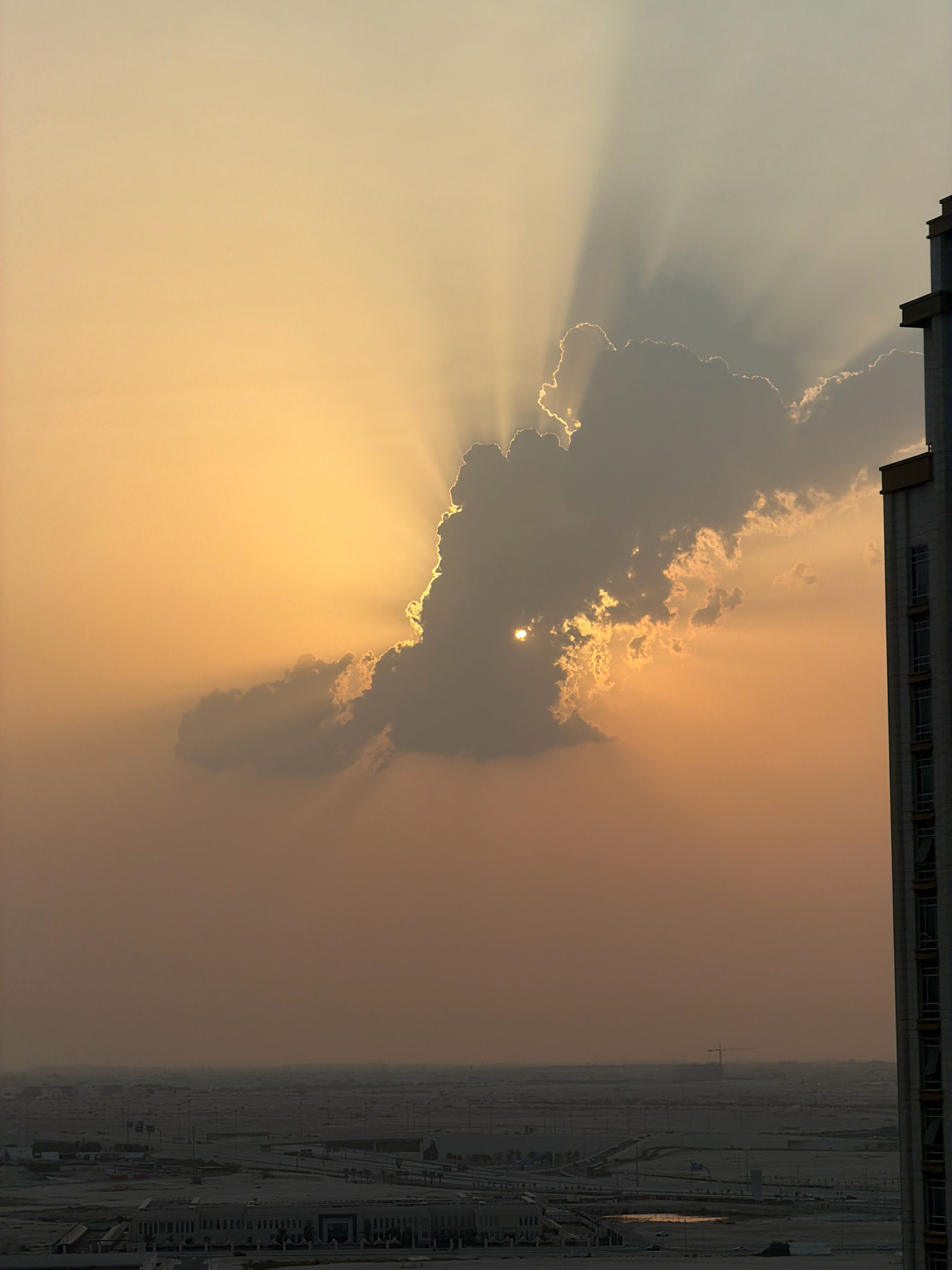 This is a vertical, eye-level, medium shot of a cityscape in Lusail, Qatar, at sunset. The sky is a gradient of warm oranges and yellows, with prominent sunbeams emanating from behind a large, dark cloud formation in the center of the frame. A portion of a tall, modern building with a facade of windows is visible on the right side of the image, extending from the top to the bottom. The lower half of the image shows a sprawling urban landscape with numerous buildings, roads, and what appears to be ongoing construction, indicated by a distant crane. The overall atmosphere is serene and highlights the dramatic interplay of light and shadow during sunset.